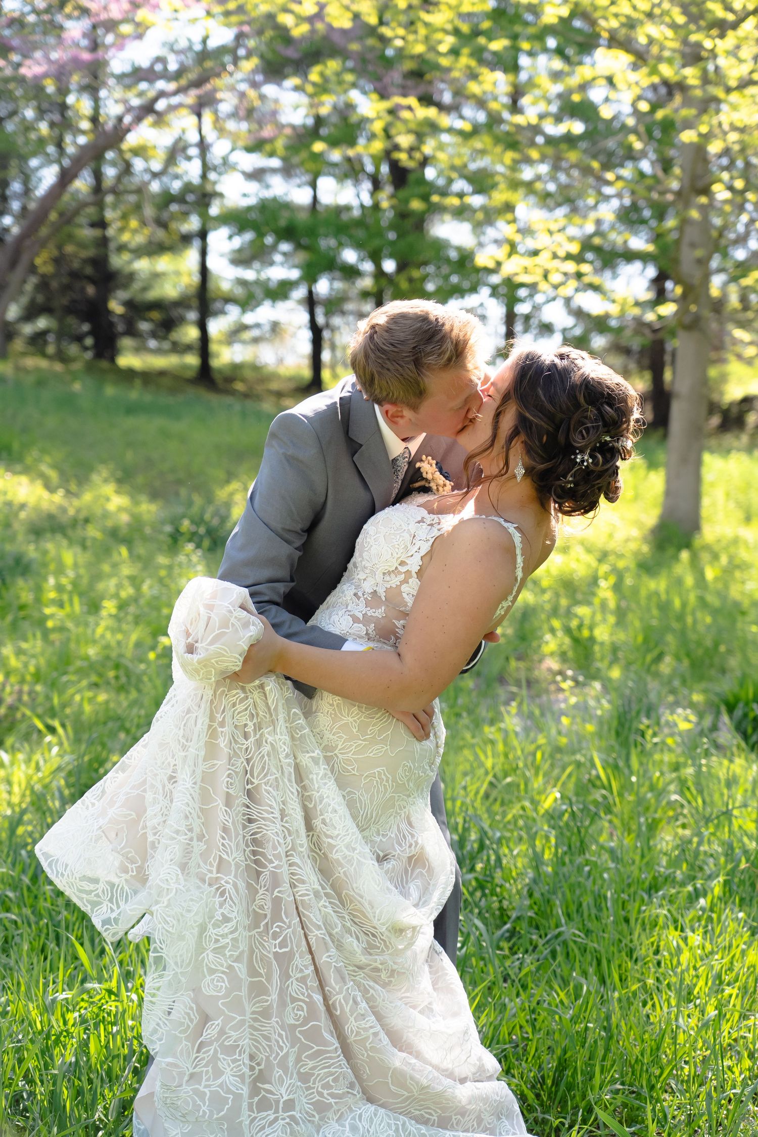 Newlyweds share a romantic kiss in a sunlit meadow with trees, the bride wearing an elegant lace wedding gown.