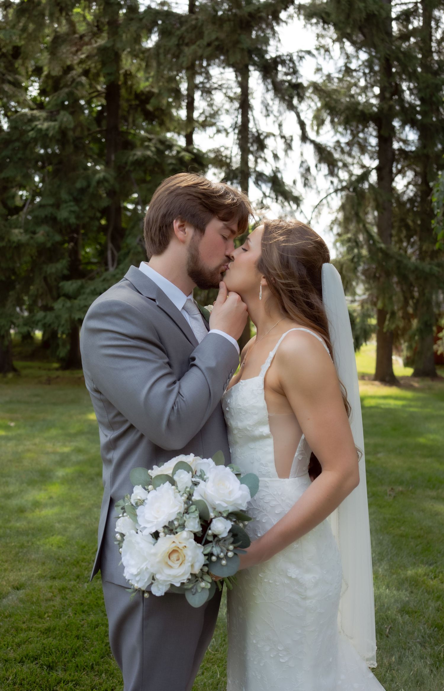 A bride in a white dress holds a bouquet of white flowers while sharing an intimate moment outdoors.