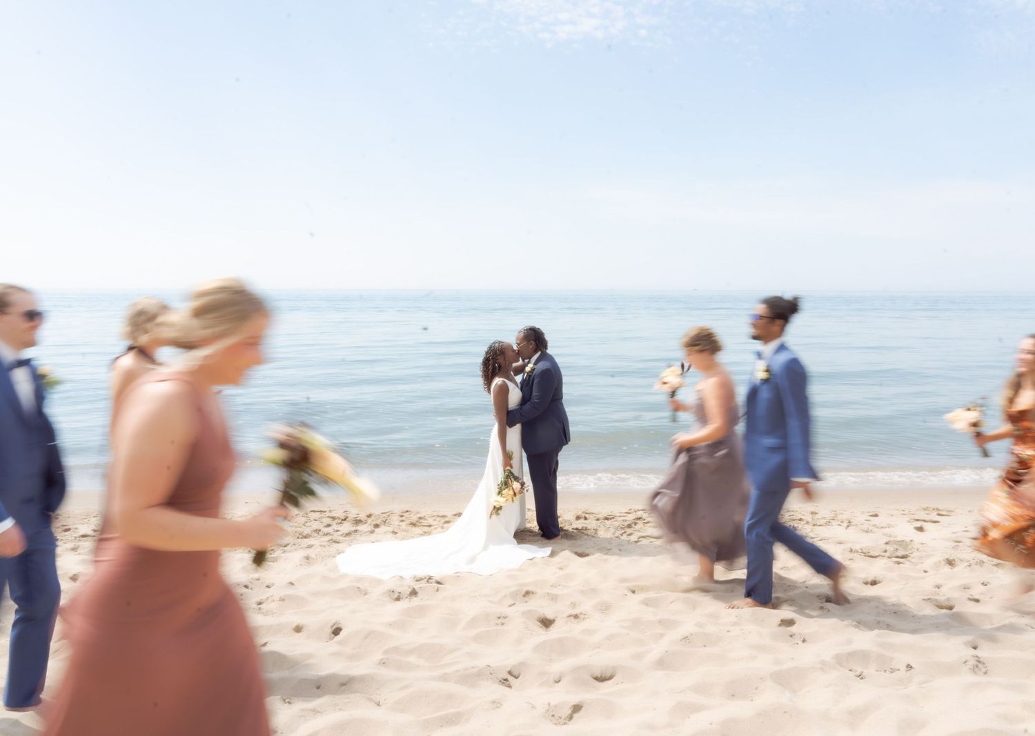 A romantic beach wedding ceremony with guests gathered on the sandy shore as couple embraces by the ocean.