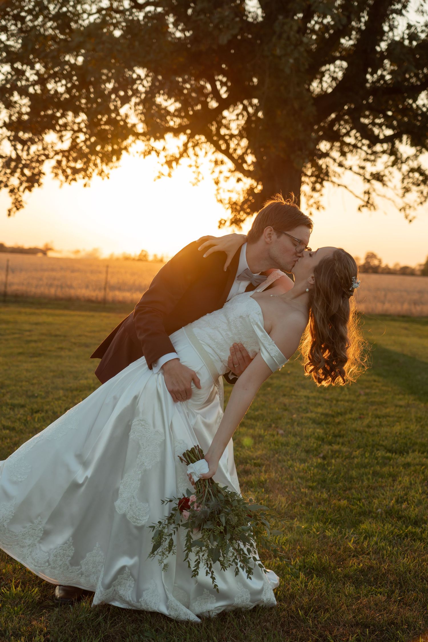A romantic sunset wedding photo with a couple in formal attire sharing a kiss in a field beneath a tree.