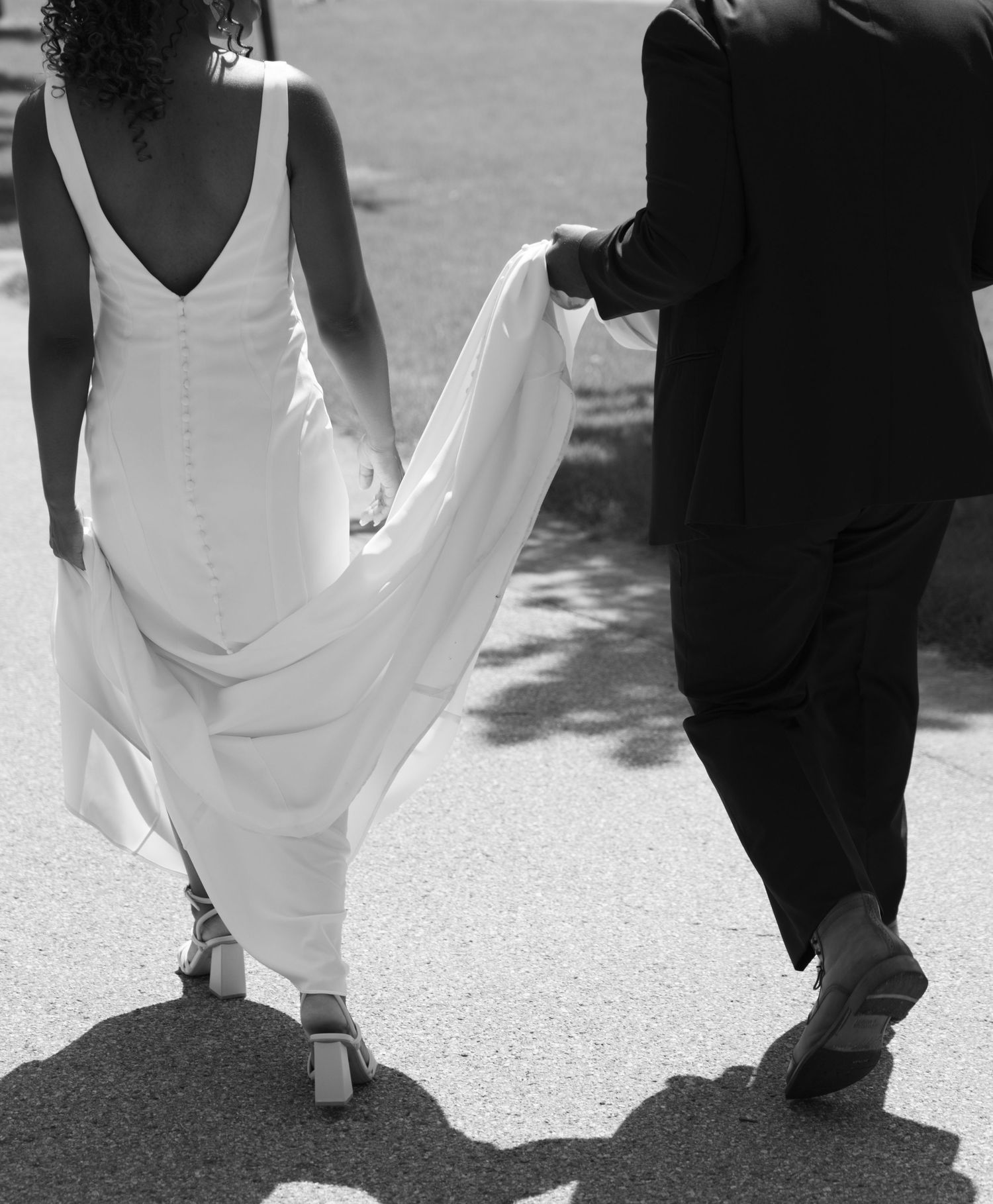 A black and white photo of a bride in a flowing white dress walking beside a person in a dark suit.