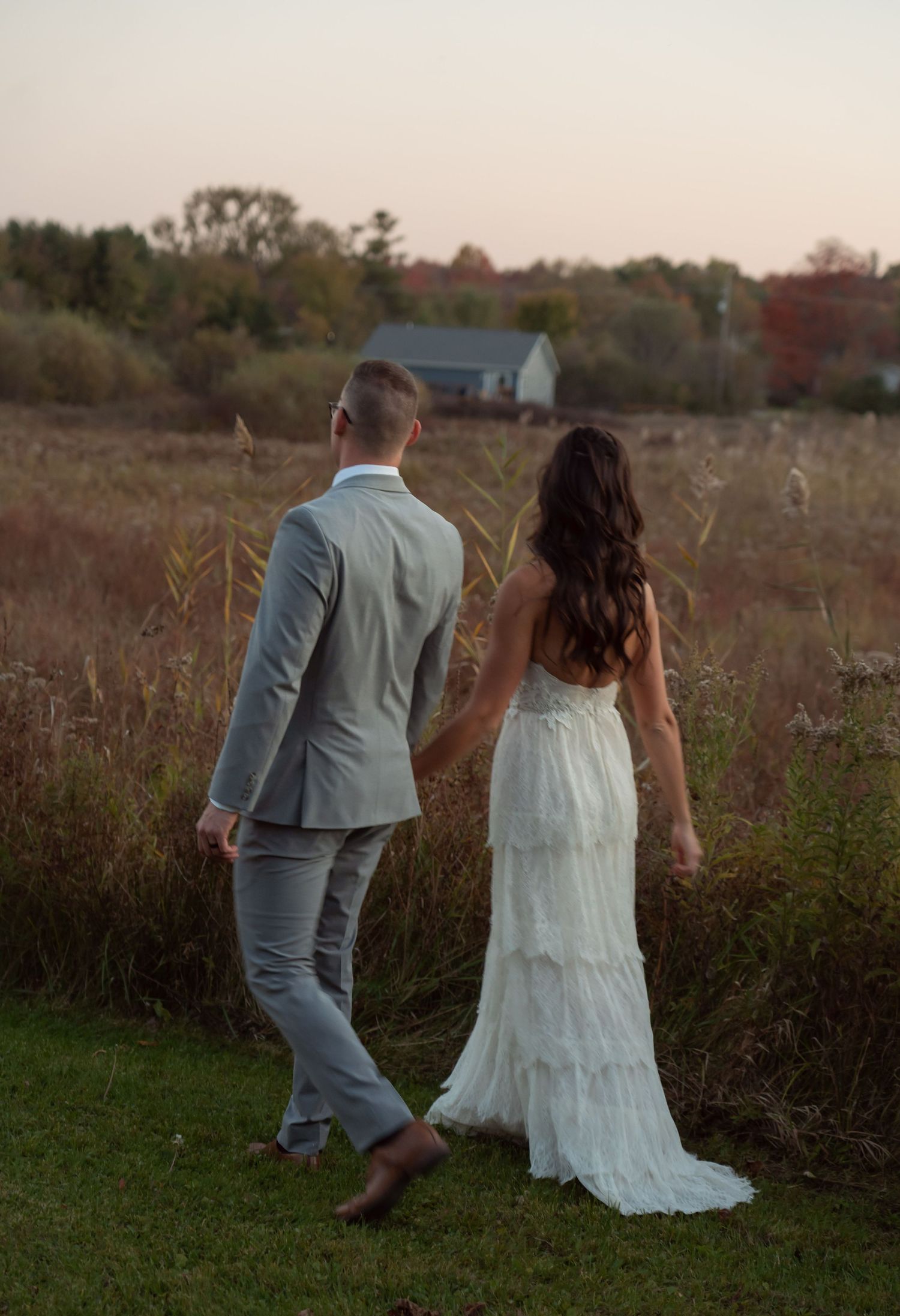 A couple in wedding attire walks through a rustic field at sunset with a small blue house in the distance.