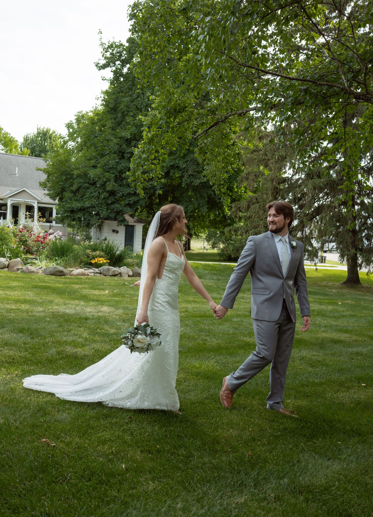 A couple in wedding attire holds hands while walking across a lush green lawn with gardens and trees in the background.