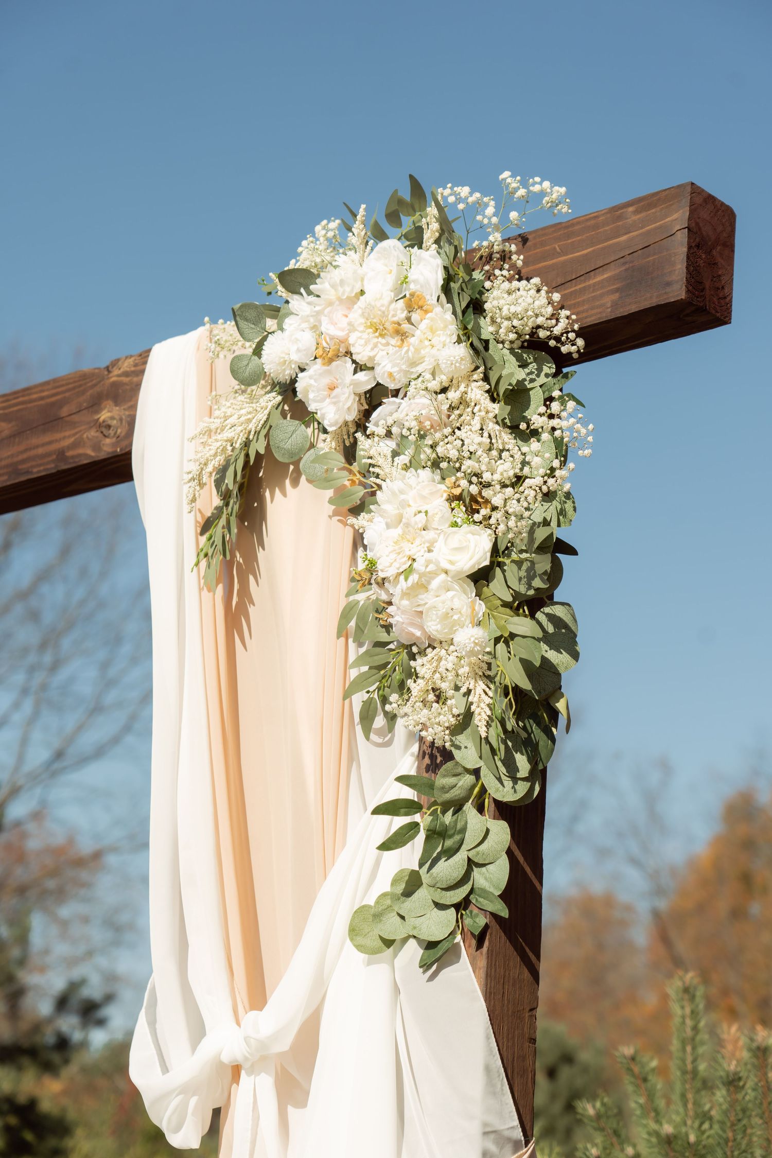 Wooden cross wedding arch decorated with white flowers, greenery and flowing fabric against blue sky backdrop.
