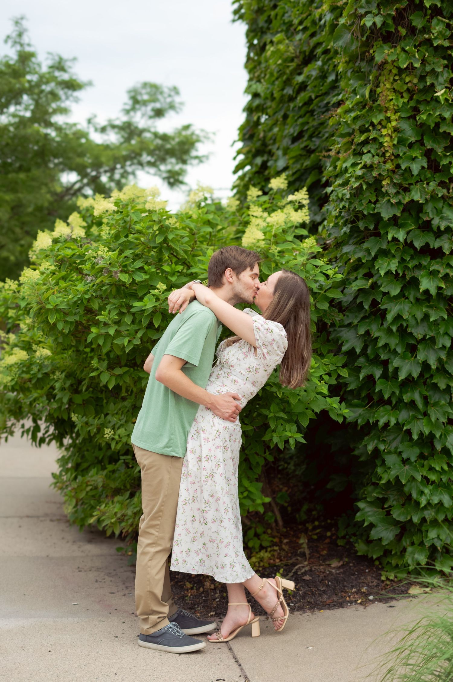 Couple embracing on a sidewalk surrounded by lush green foliage.