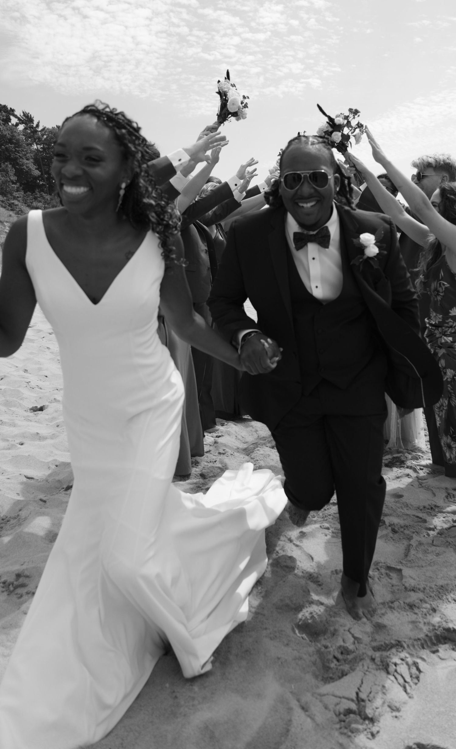 Joyful newlyweds celebrate on a beach with guests raising their hands in celebration during a sunny outdoor wedding.
