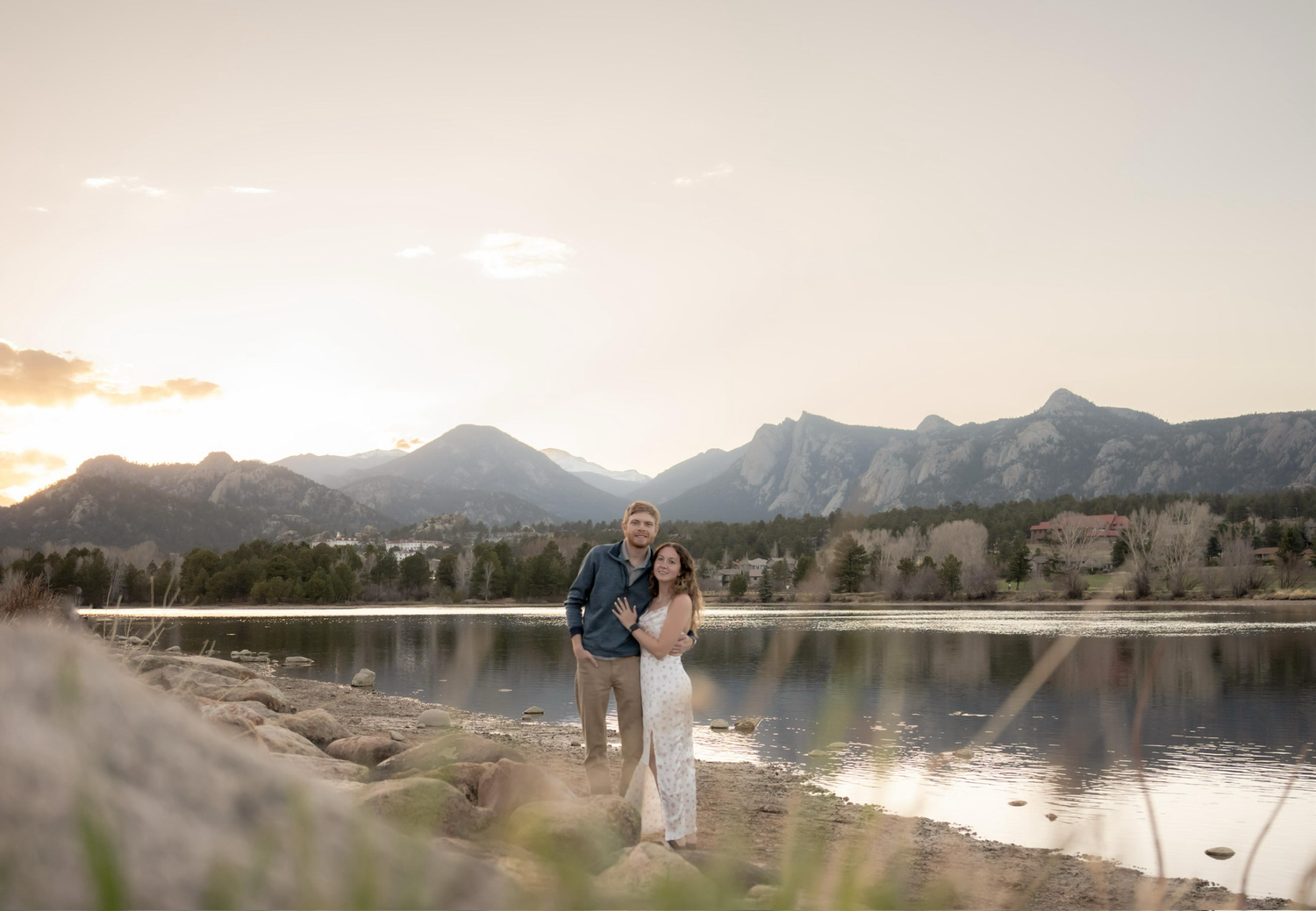 A couple stands near a serene mountain lake at sunset, surrounded by majestic peaks and natural landscape.