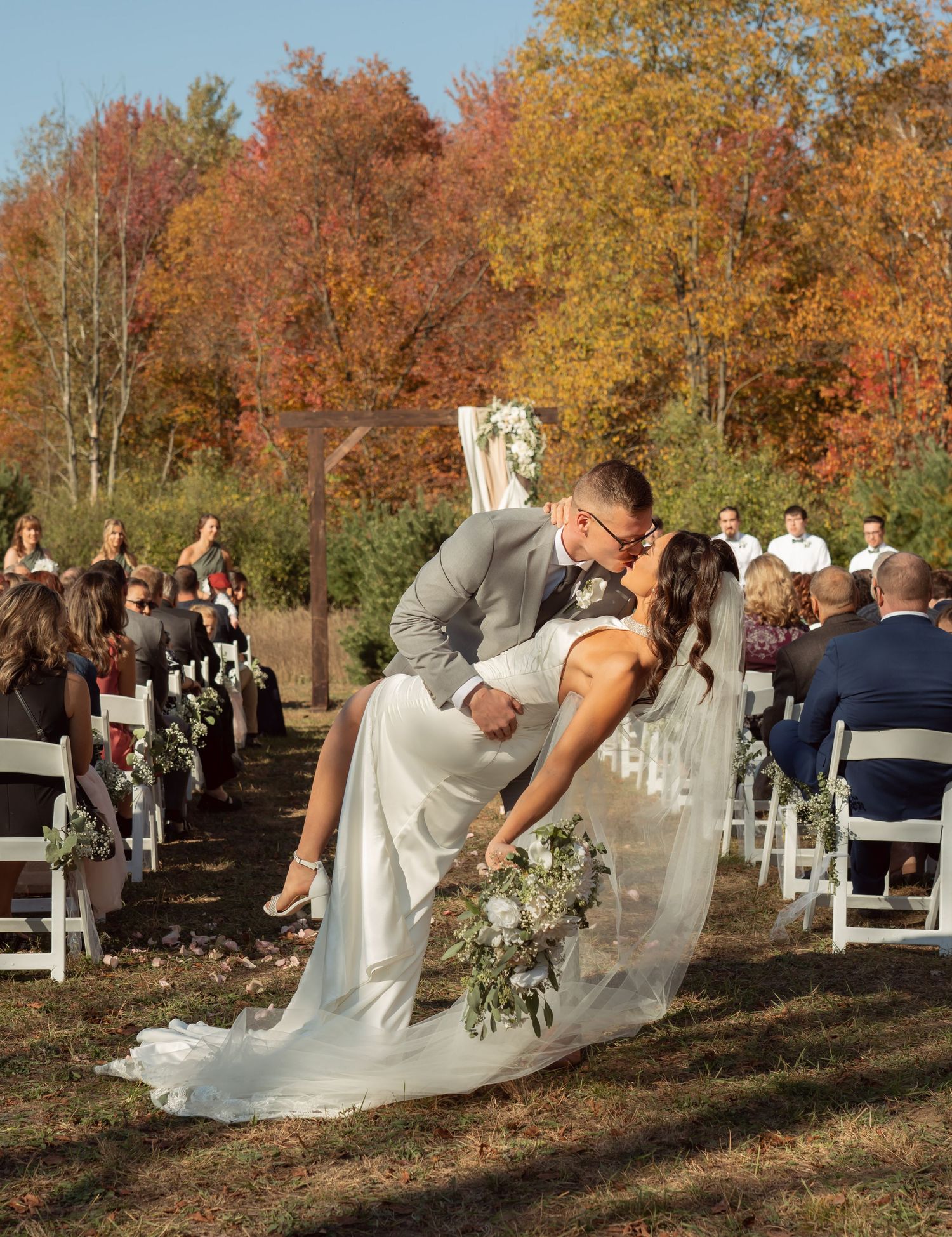 A romantic wedding ceremony takes place outdoors with couples dancing amid vibrant autumn foliage and white chairs.