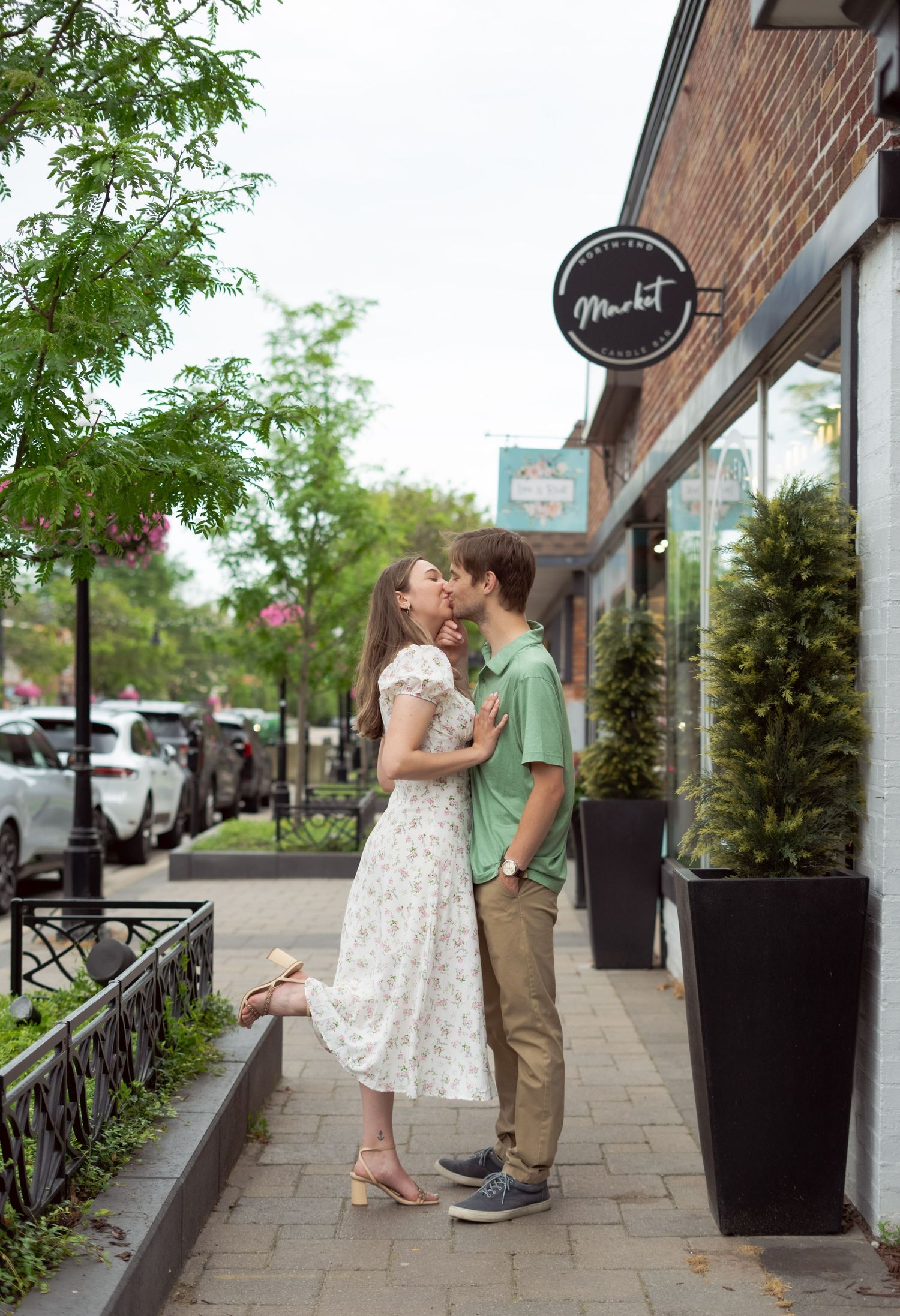 A romantic moment shared between a couple in front of a Market storefront on a tree-lined city sidewalk.