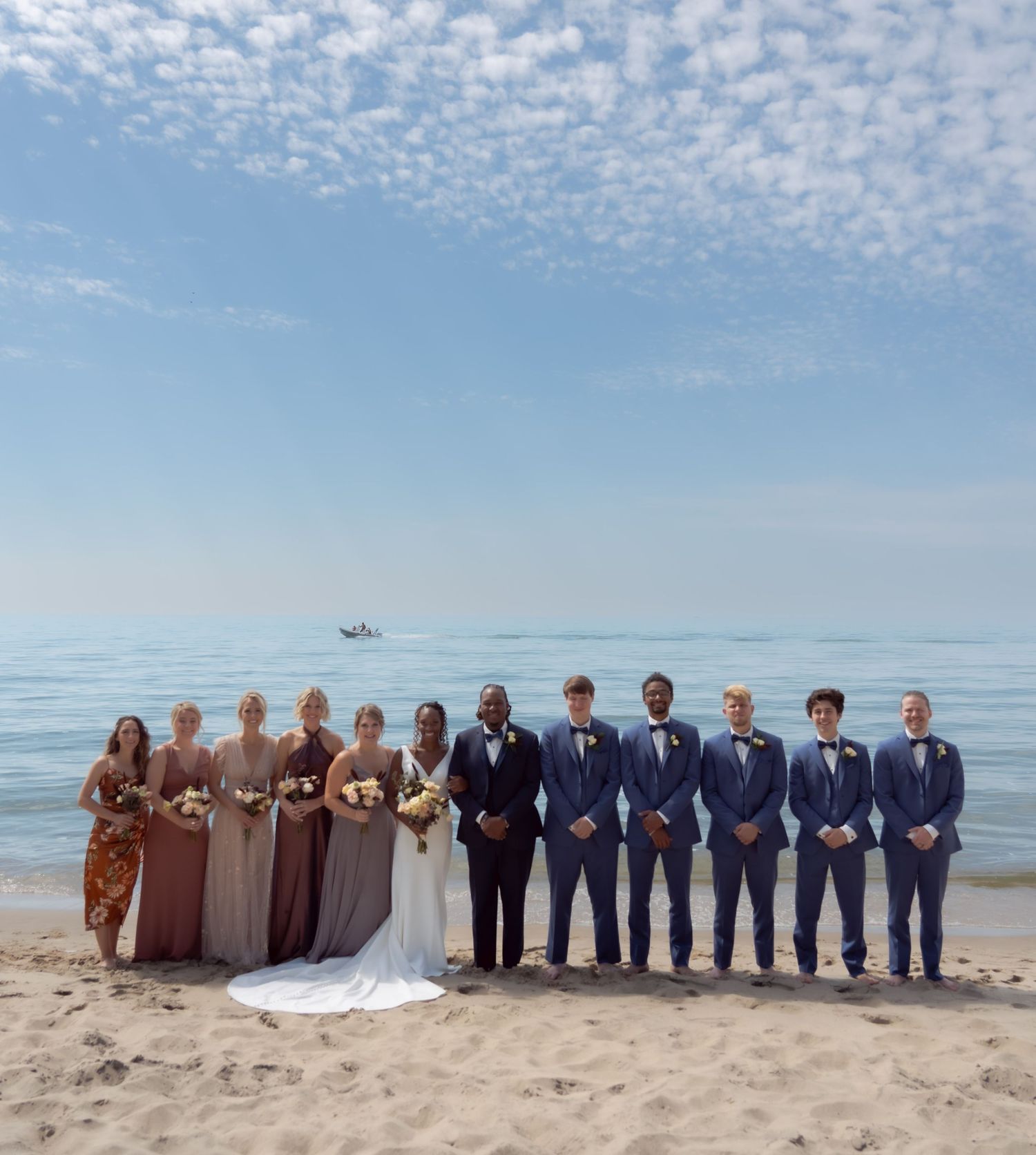 Wedding party stands lined up on sandy beach with cloudy blue sky and calm ocean in the background.