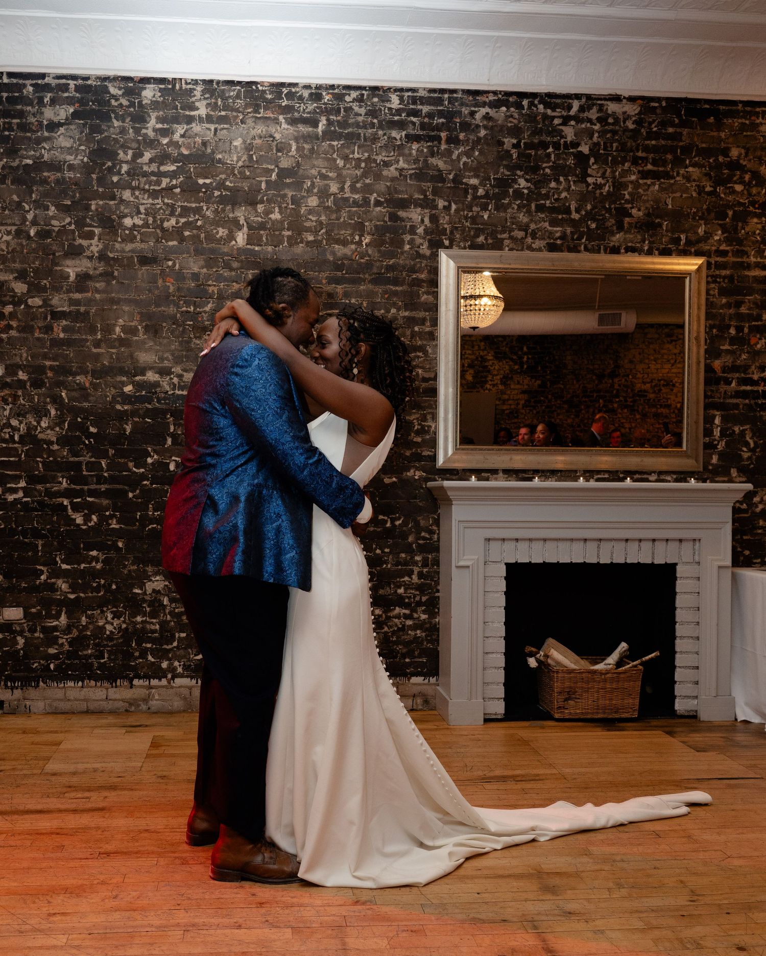 A couple shares their first dance in formal attire against a rustic brick wall with a fireplace in an intimate wedding venue.