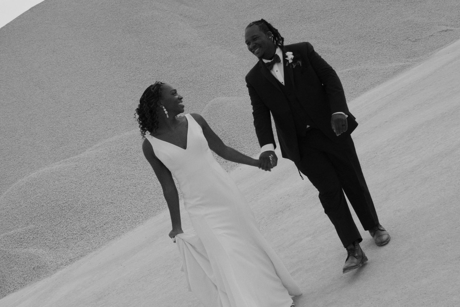 A bride and groom hold hands while walking together in a romantic black and white outdoor wedding photo.