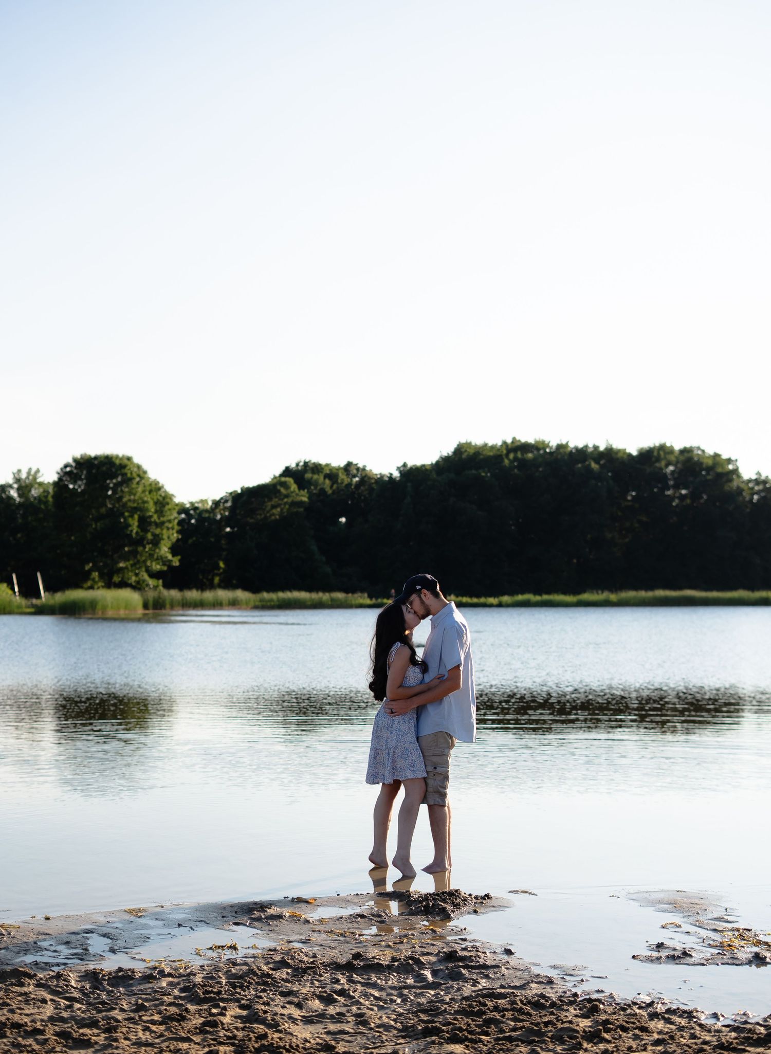 Two people standing on a rocky shoreline by a tranquil lake surrounded by trees during a bright summer day.