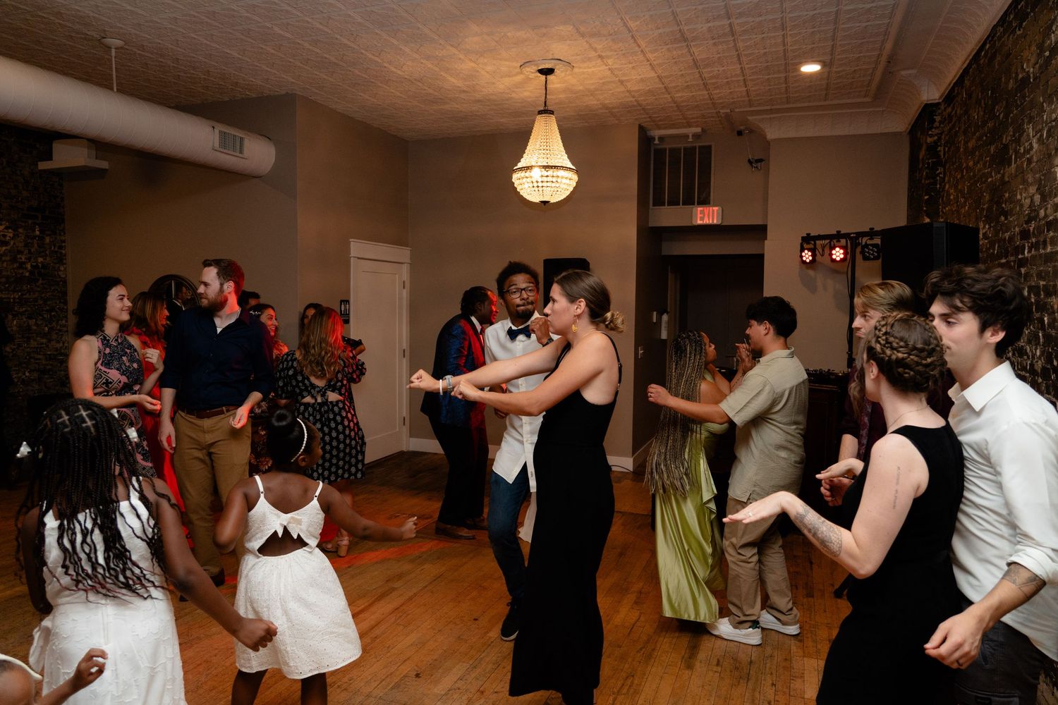 Couples dance together on a wooden floor under a chandelier at an indoor evening social event.