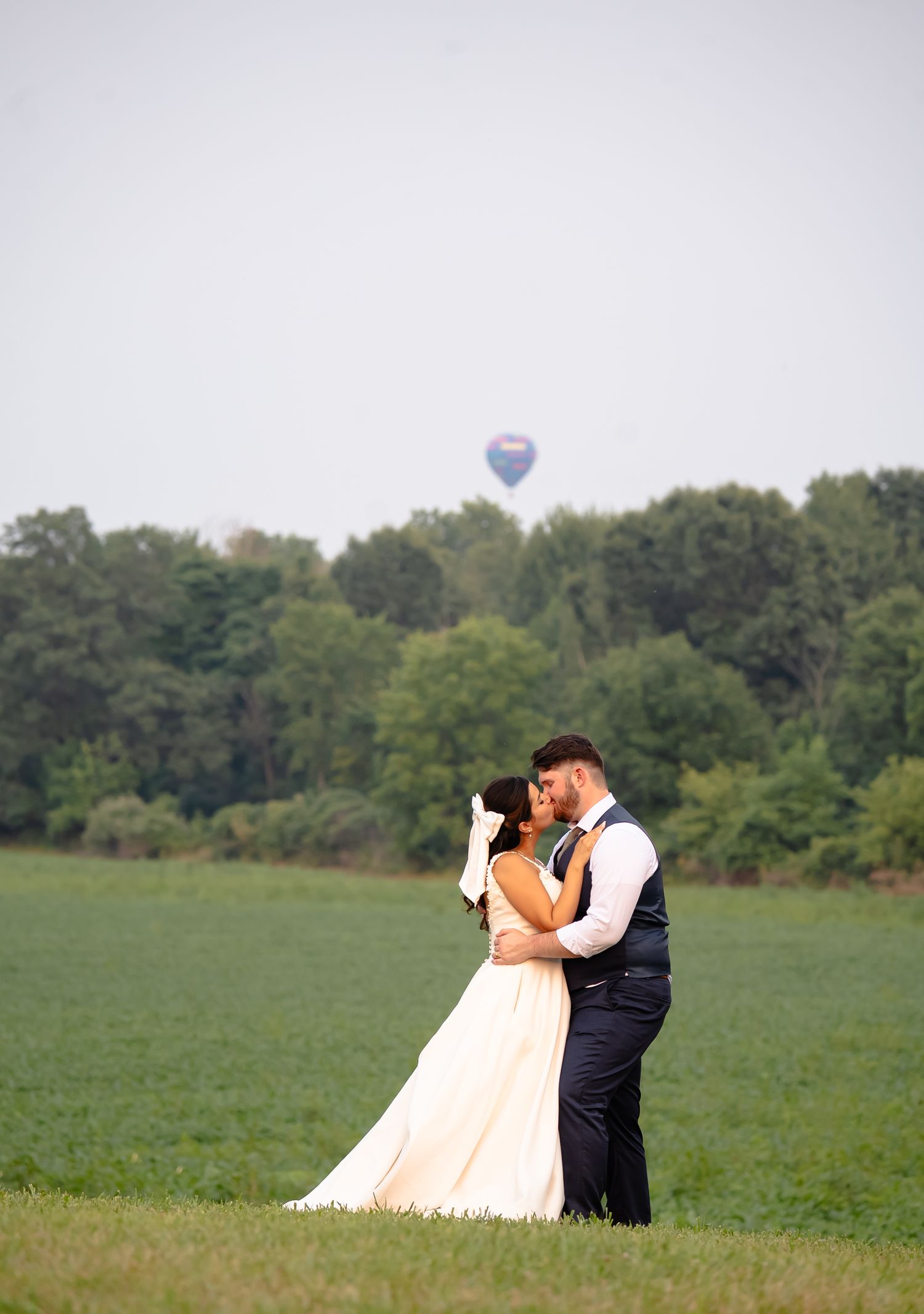 A couple in wedding attire embraces in a green field with a hot air balloon floating in the background.