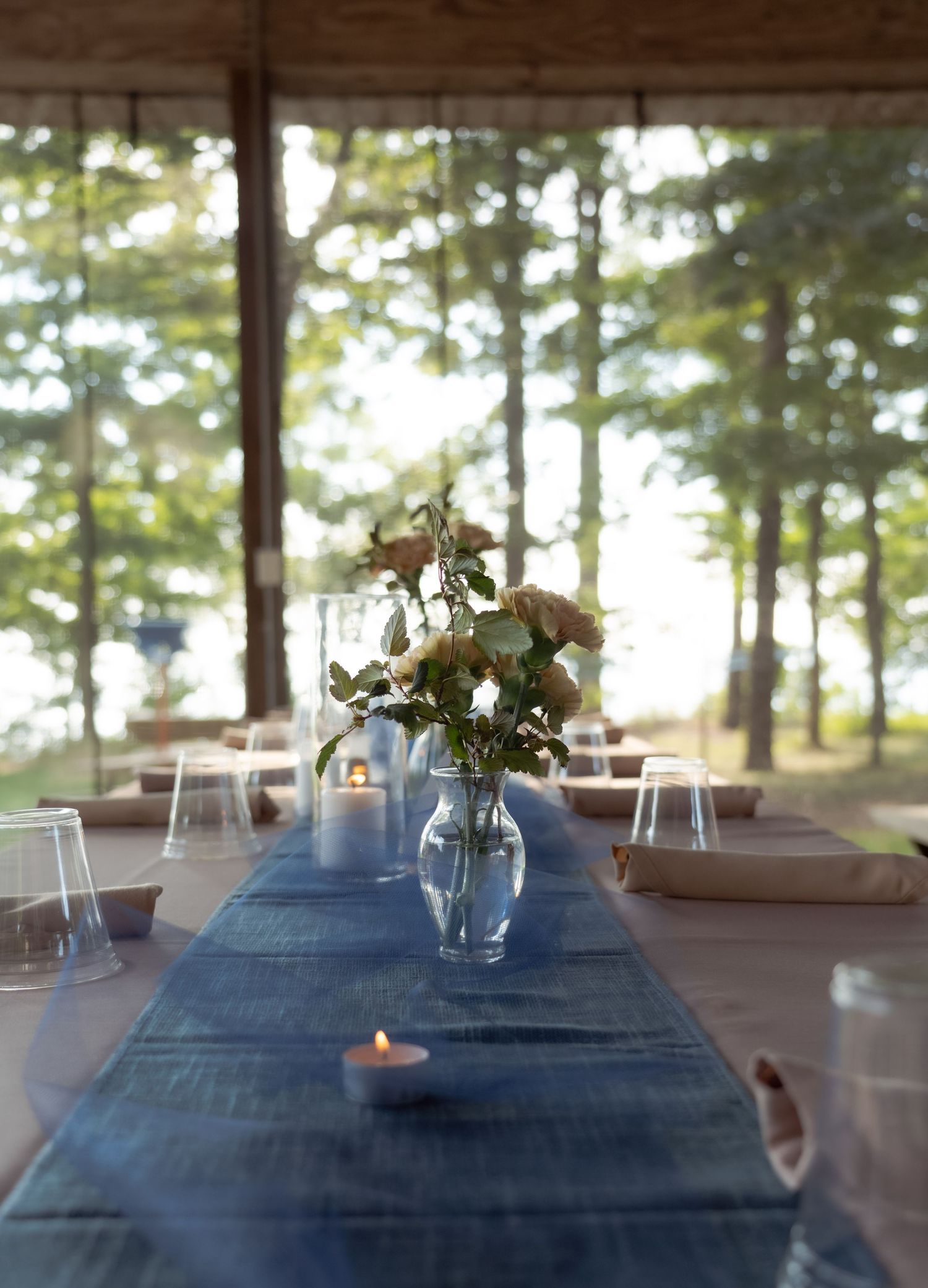 Rustic outdoor wedding table with blue runner, floral centerpiece, and forest backdrop.