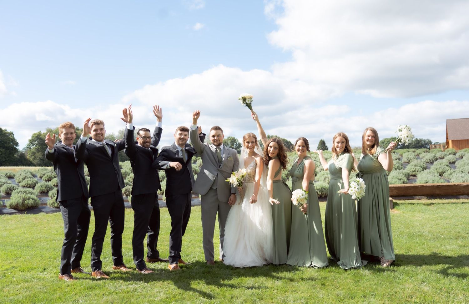 Wedding party celebrates outdoors on a sunny day with groomsmen in black suits and bridesmaids in sage green dresses.