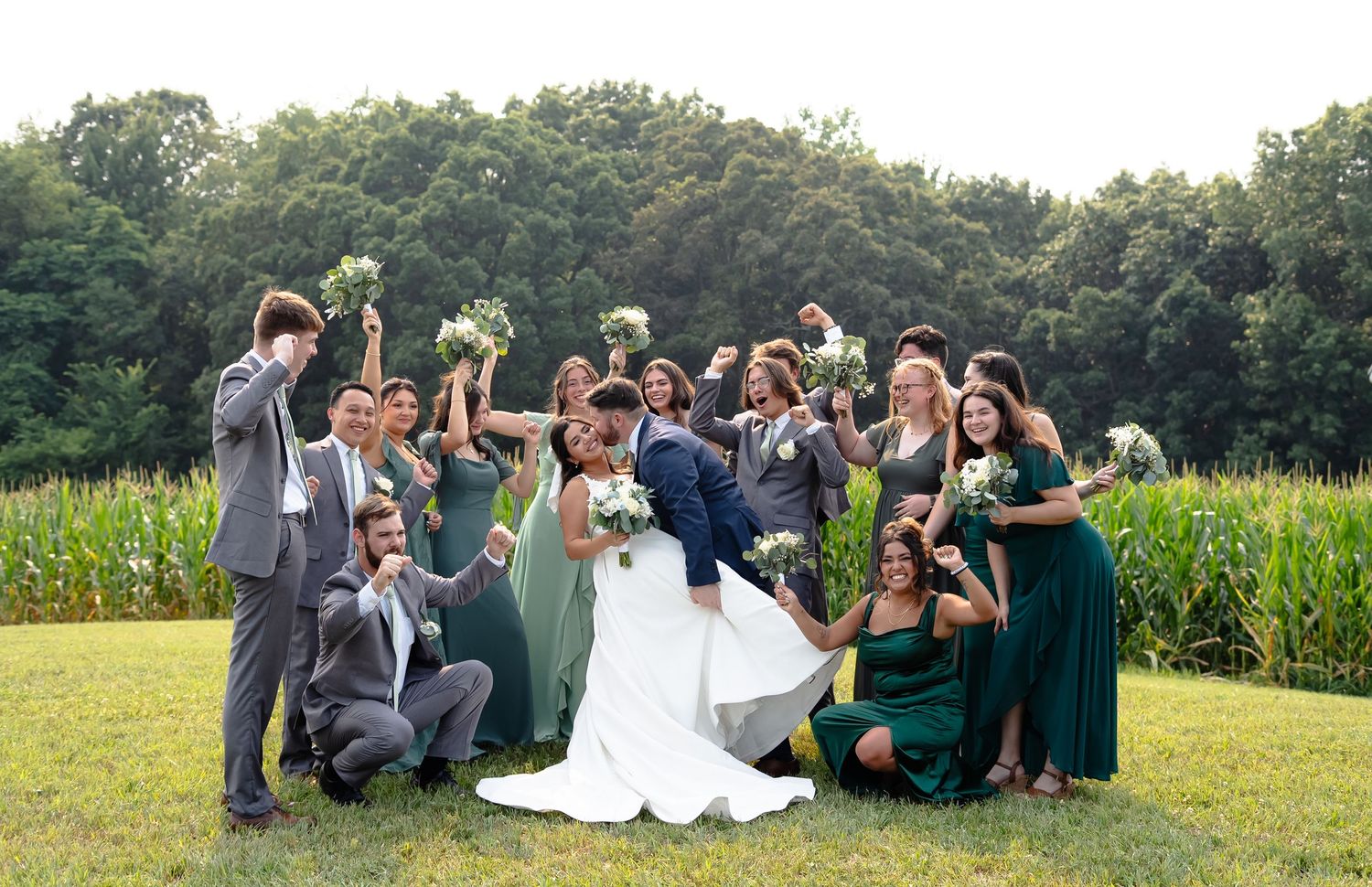 Wedding party sits together on grassy lawn during outdoor summer celebration in front of cornfield.