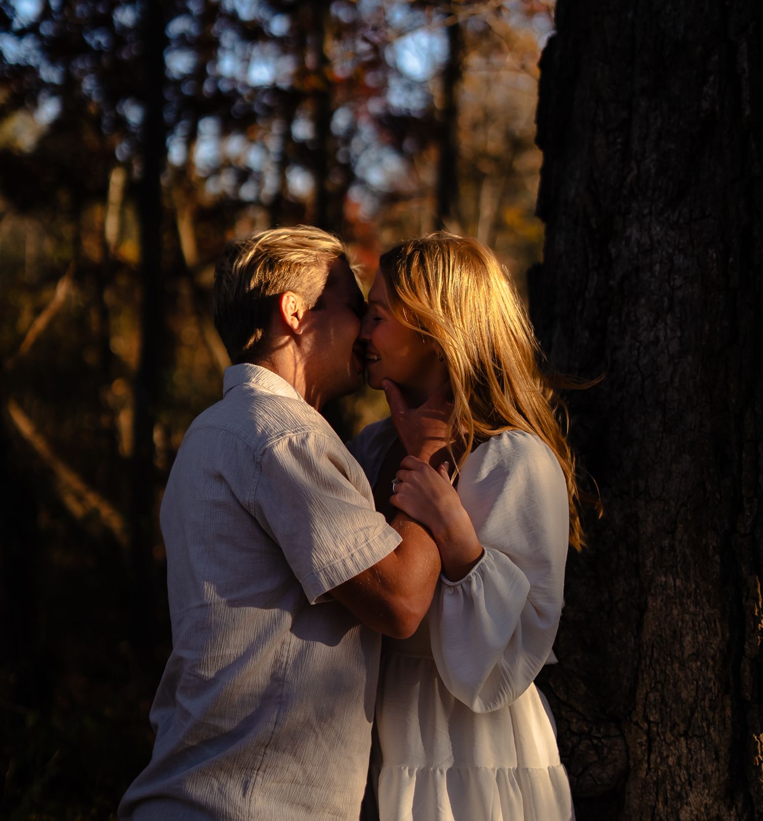 A romantic couple in white clothing embraces during golden hour sunset in a wooded outdoor setting.
