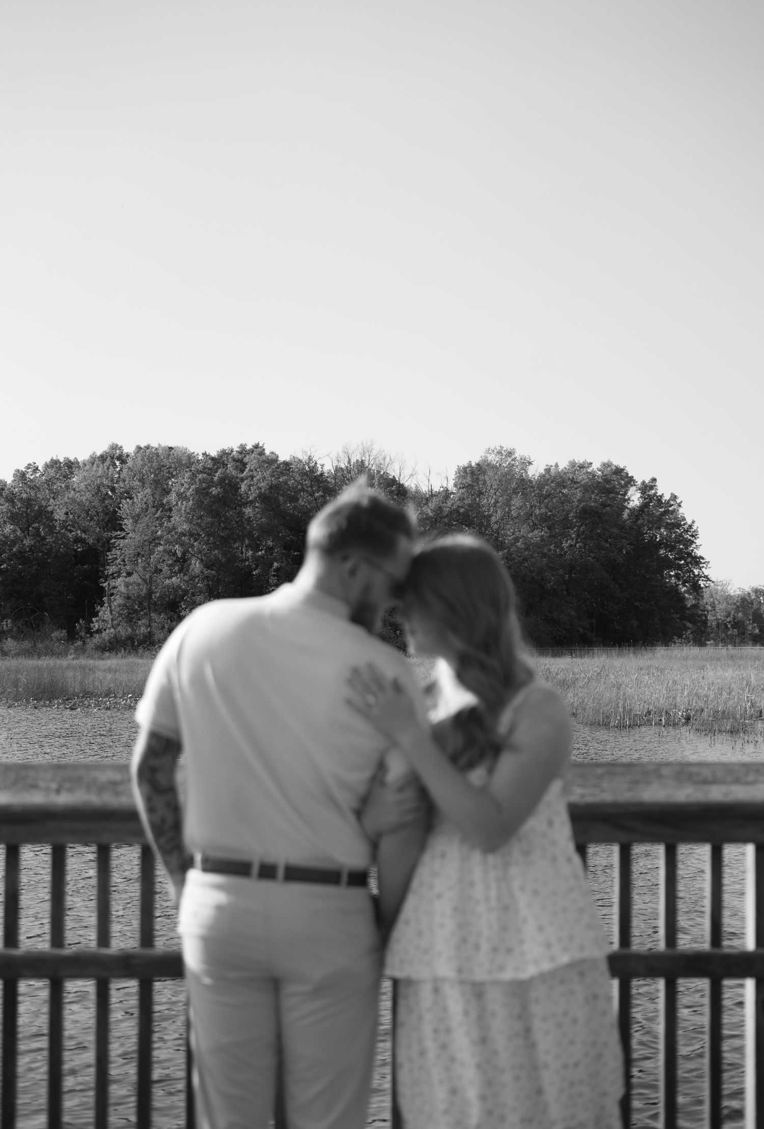 A tender black and white romantic moment shared between a couple on a wooden deck overlooking a lake and treeline.