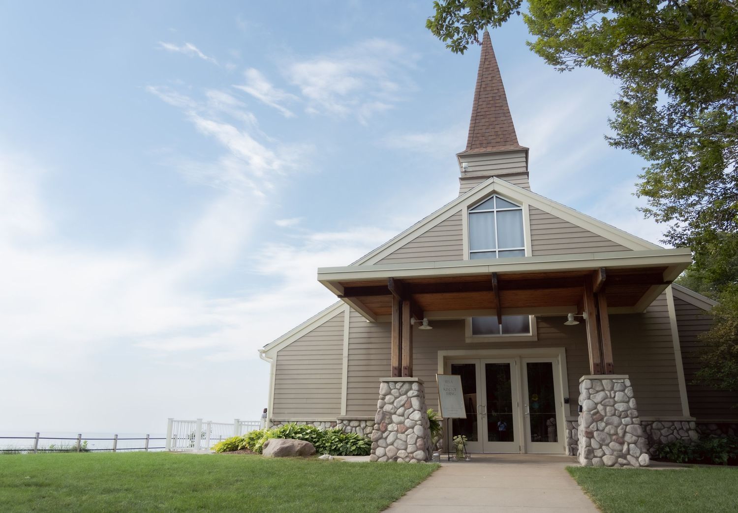 Small tan chapel with stone pillars and steeple sits on grassy lawn under blue sky at waterfront location.