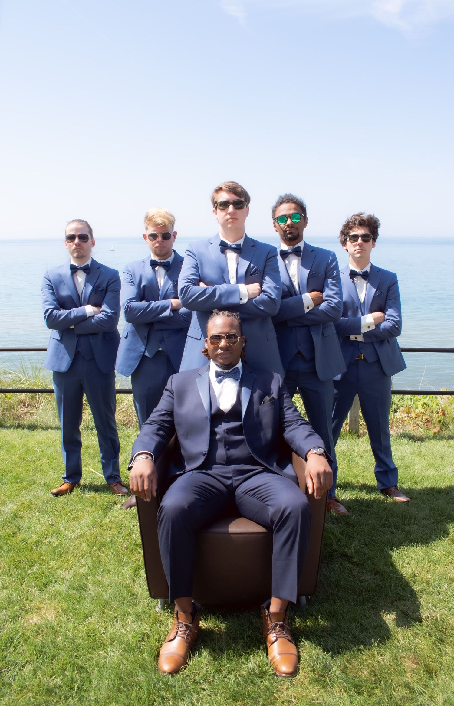 Groomsmen in matching navy blue suits pose outdoors by the ocean with one member seated in a leather chair.