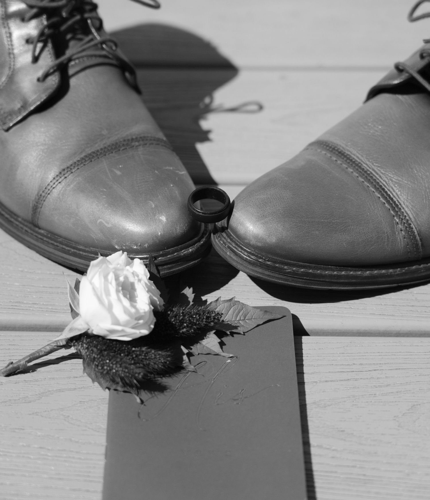 A black and white photograph shows a pair of worn leather dress shoes with a withered white rose between them.