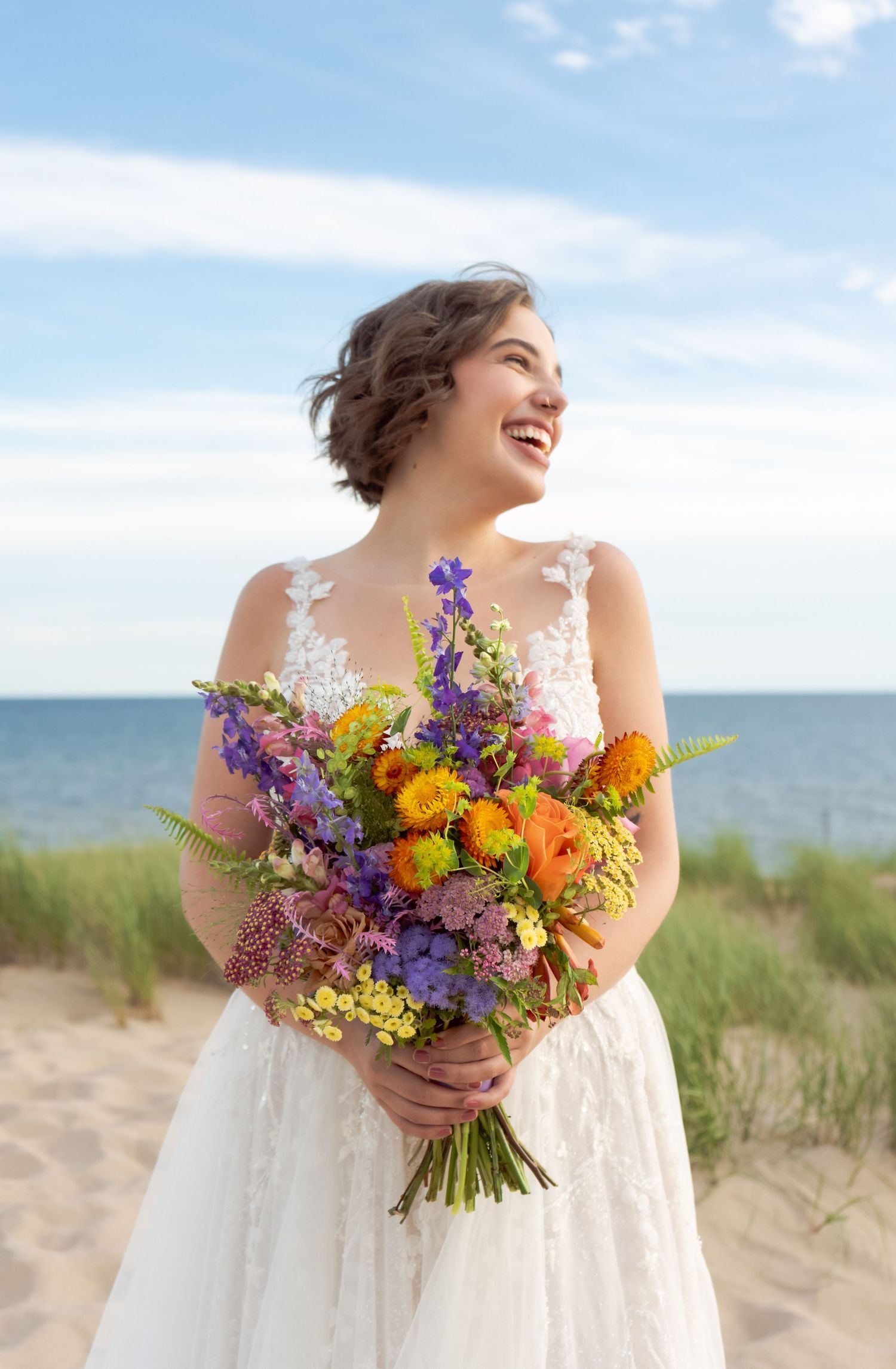 A joyful bride holding a vibrant wildflower bouquet of purple and orange blooms on a sunny beach.