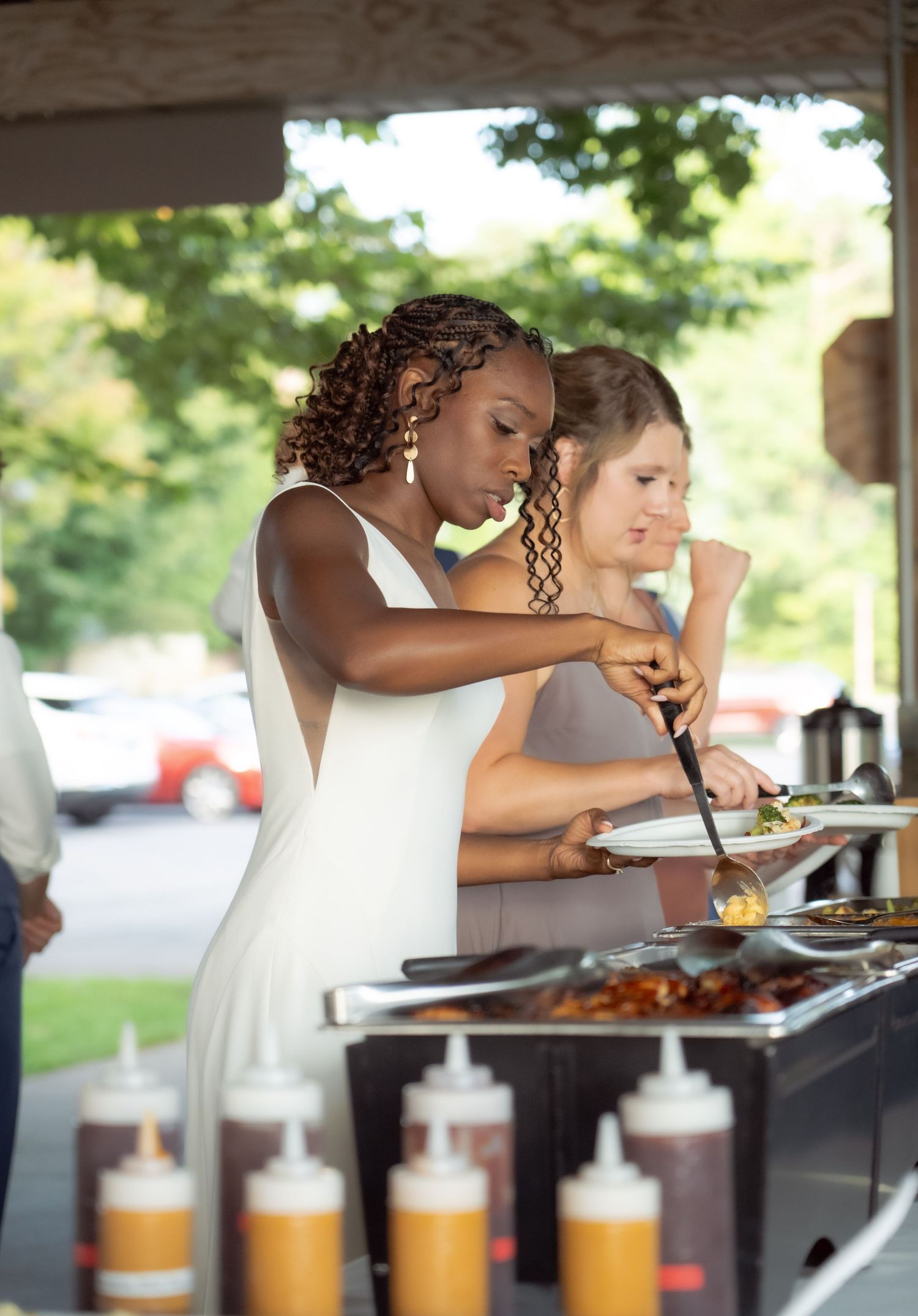 Two people in white dresses serve themselves food at a buffet line with condiment bottles visible in the foreground.