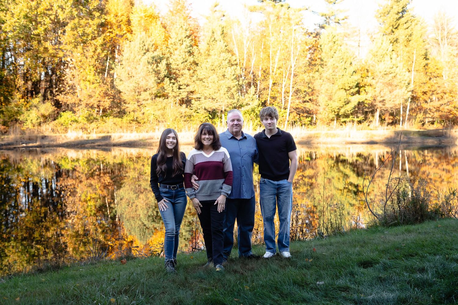 A group stands together on grass by a serene pond with golden autumn trees reflecting in the still water.