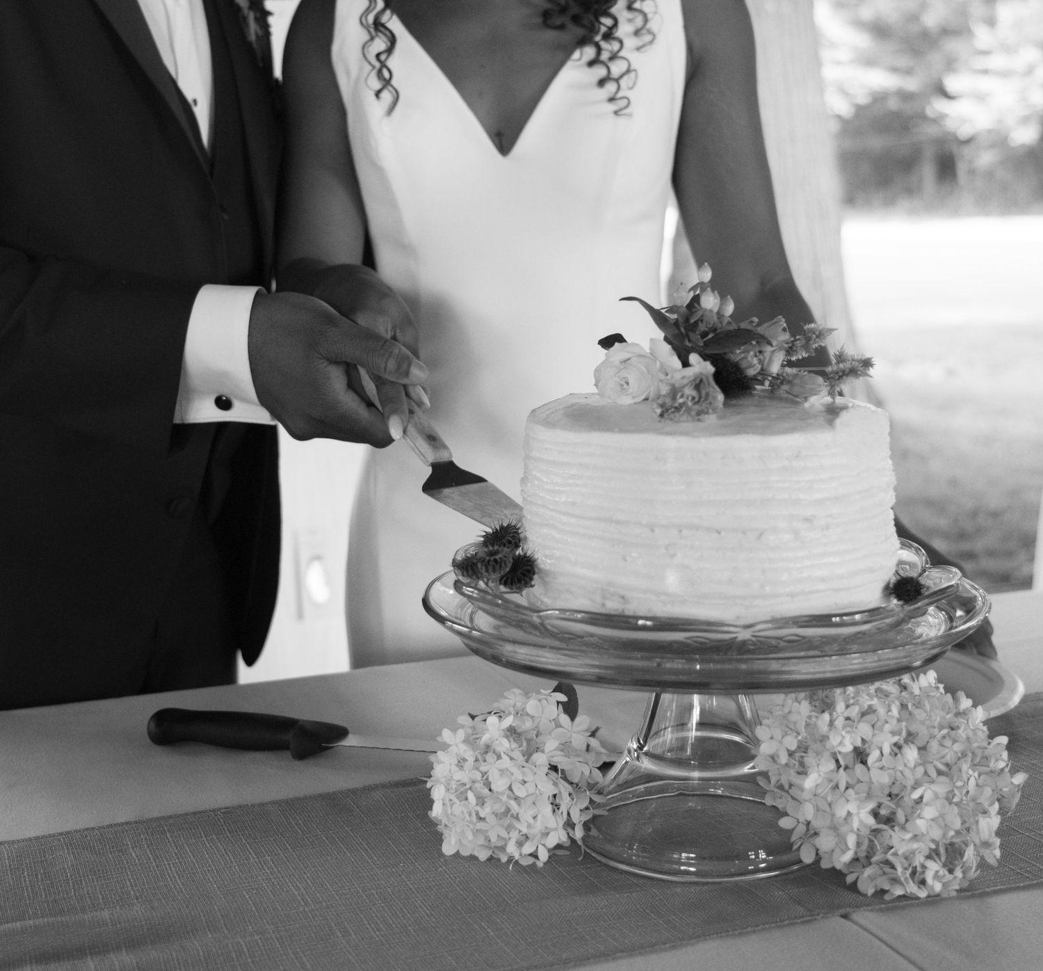 Black and white photo of a wedding couple cutting their white tiered cake decorated with flowers and set on a glass stand.