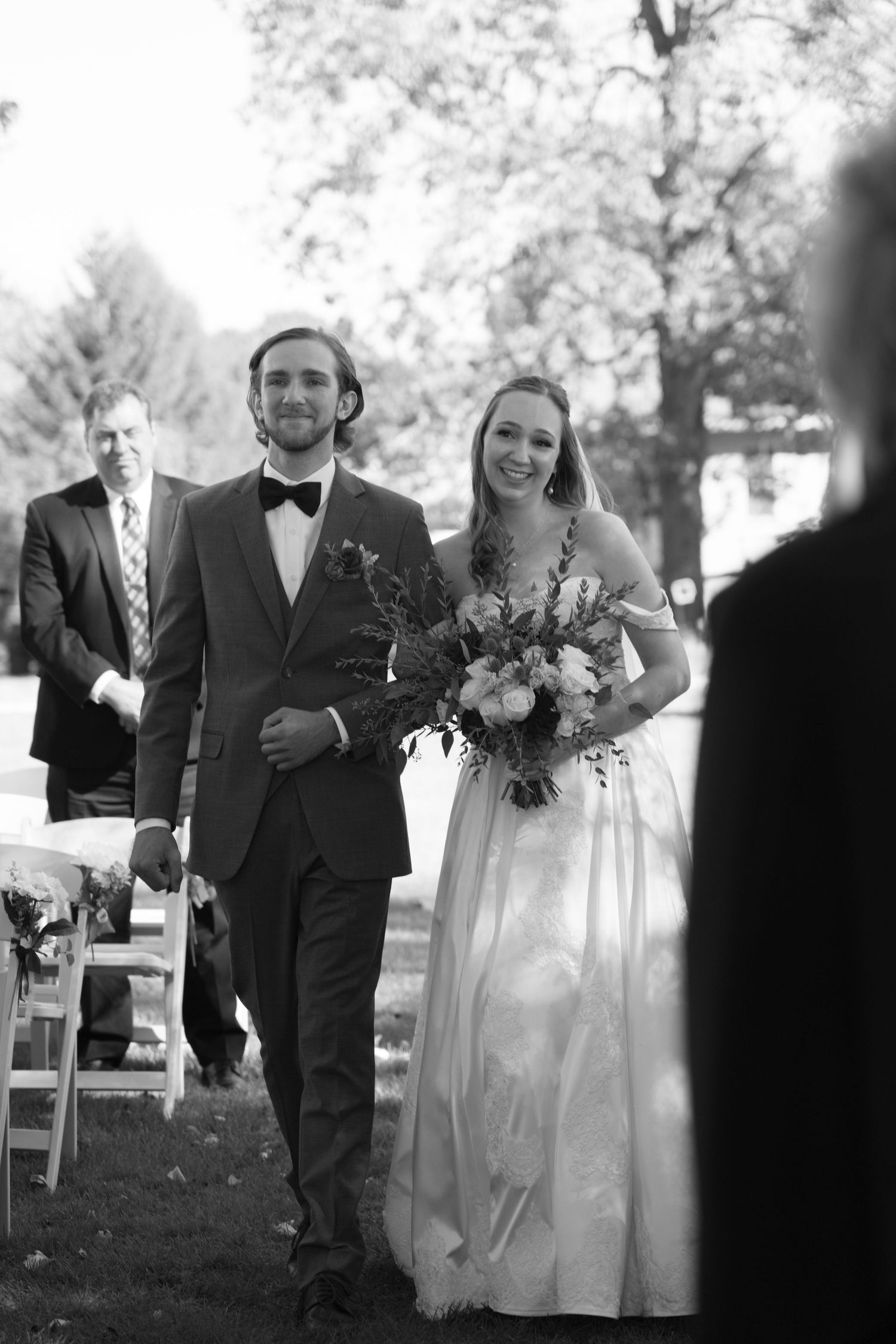 Black and white photo of couple walking down aisle during outdoor ceremony.