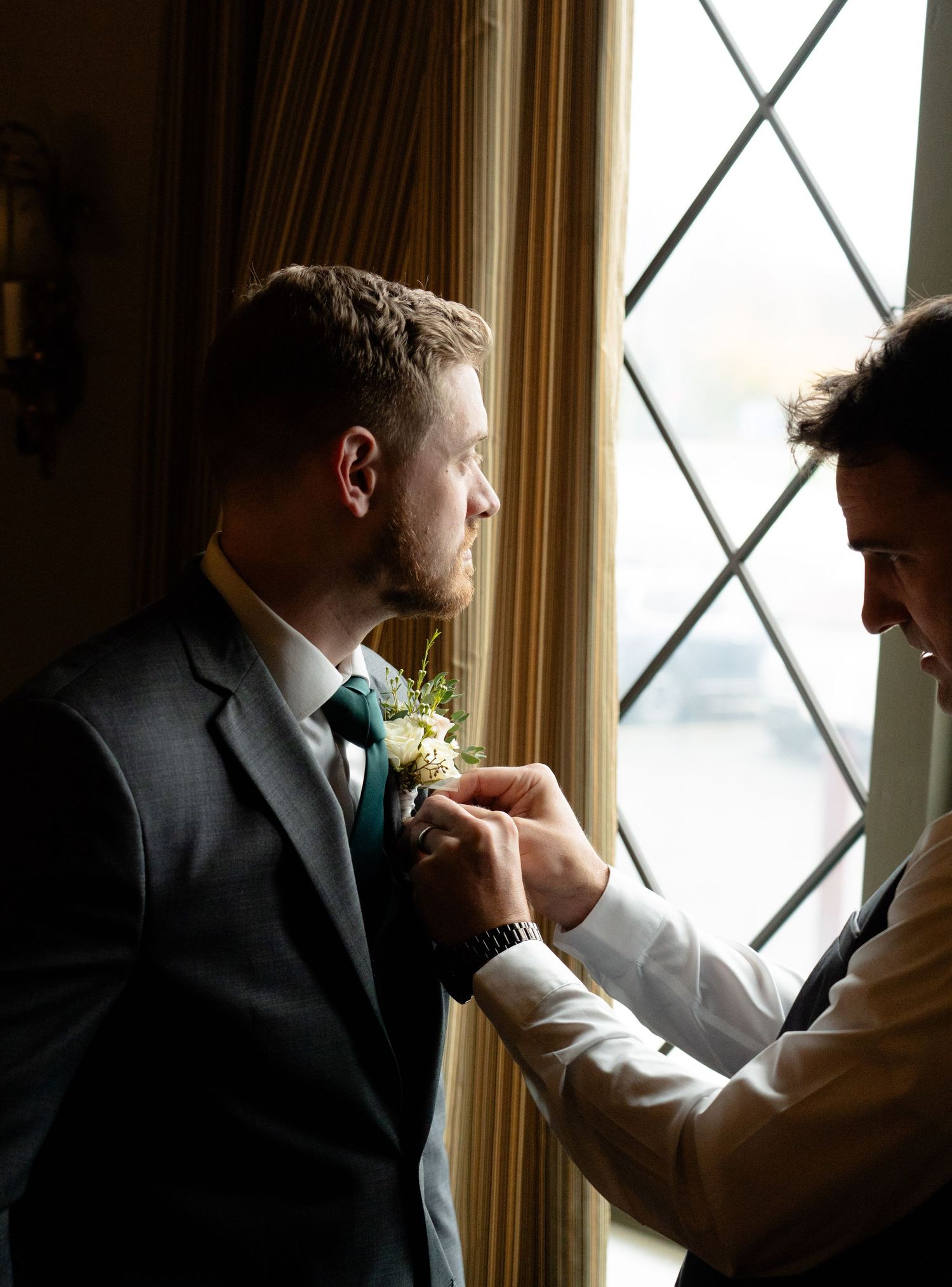 Close-up of a boutonniere being pinned onto a groom's suit jacket near a window.