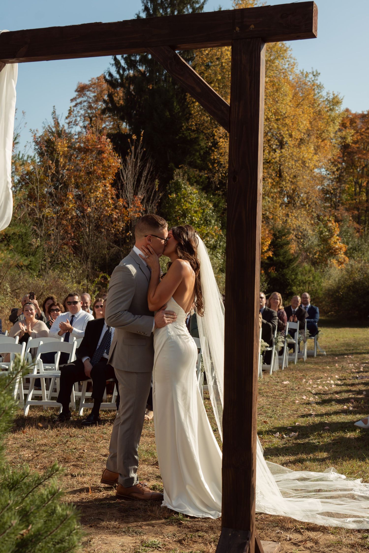 A couple shares a kiss under a wooden arch during an autumn outdoor wedding ceremony as guests look on from white chairs.