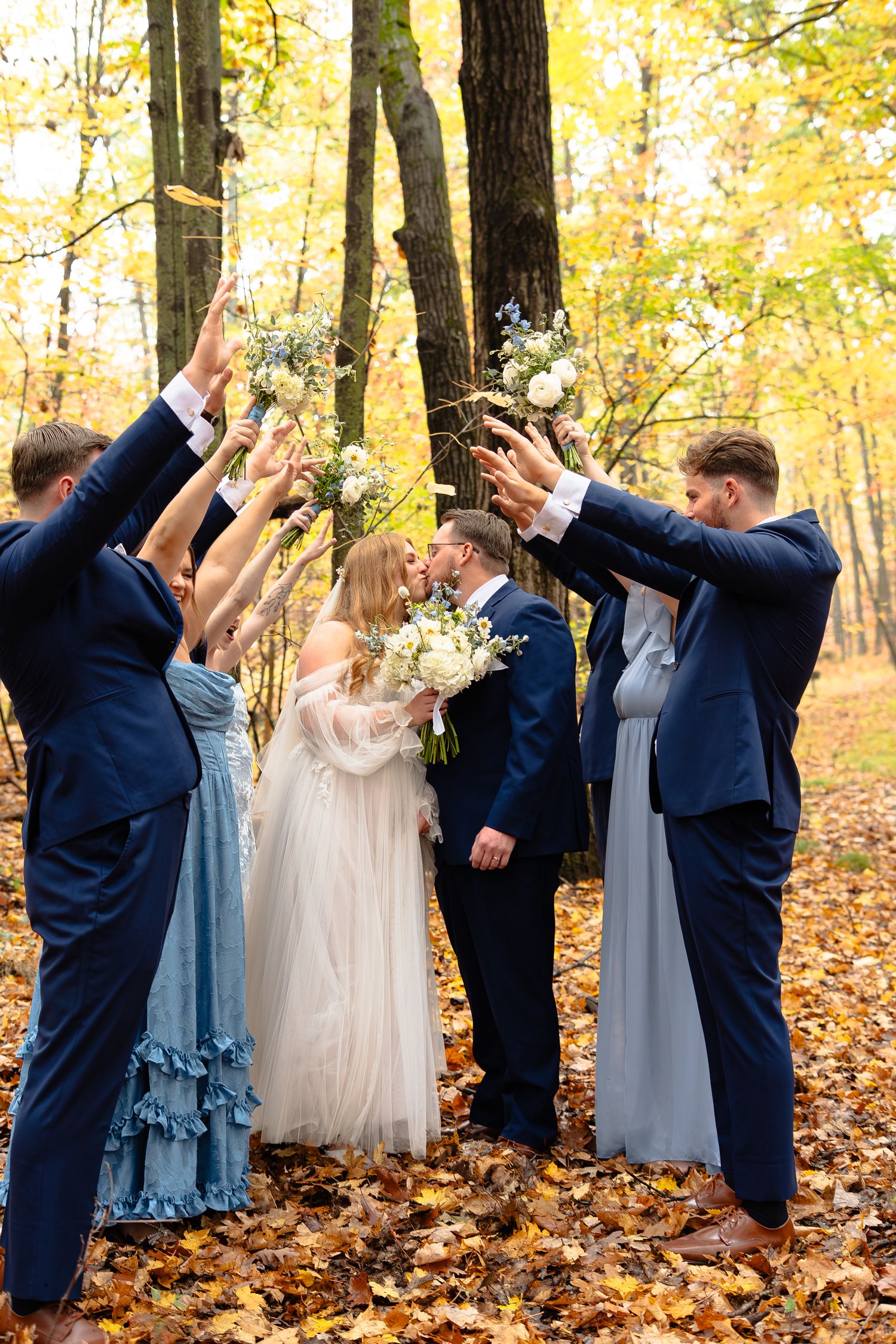 Wedding party in navy suits and light dresses pose playfully dabbing in a forest filled with golden autumn leaves.