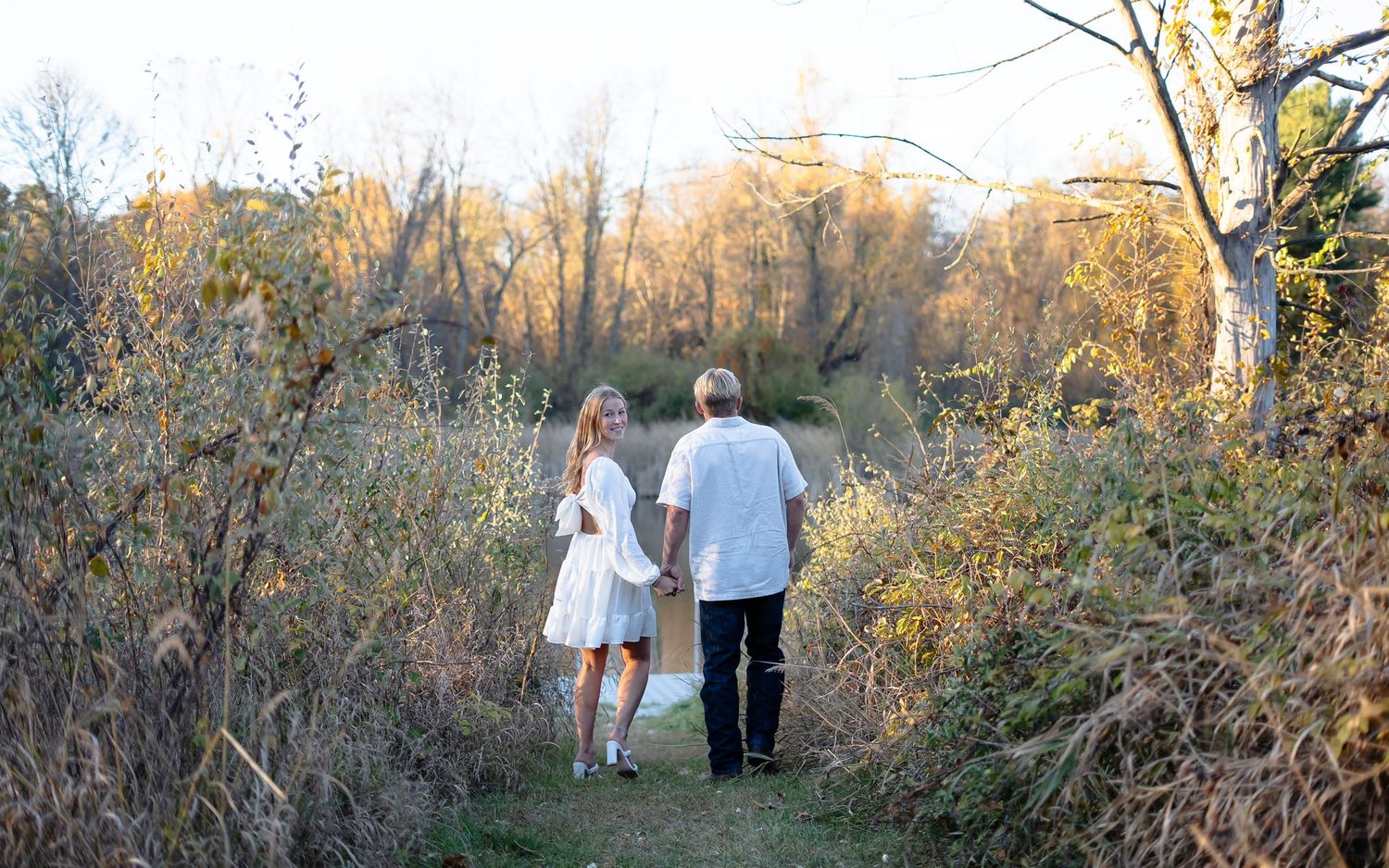 Couple walking together on a nature trail during golden hour with autumn trees in the background.