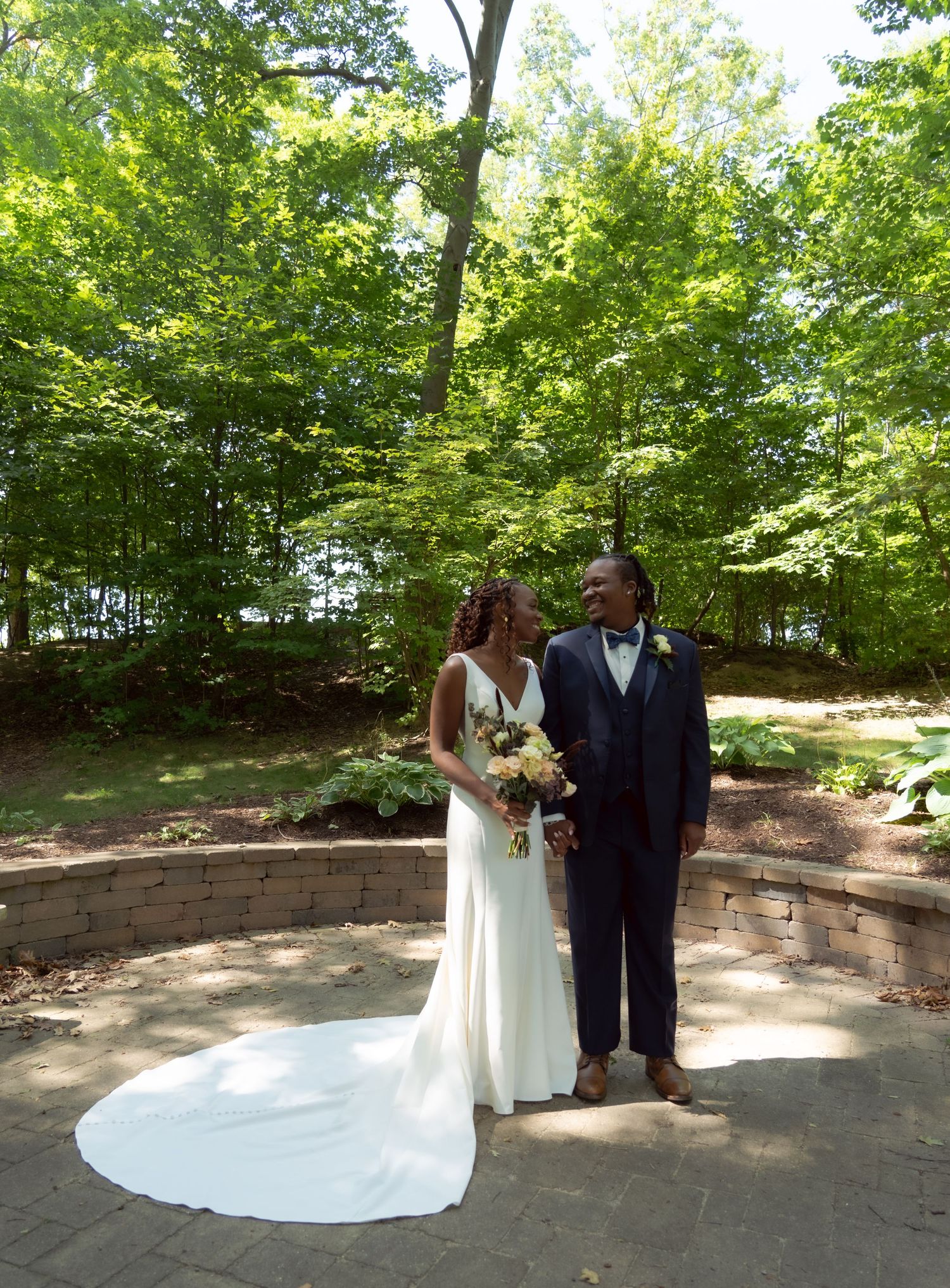 A couple standing together on a stone patio surrounded by lush green trees during an outdoor wedding ceremony.