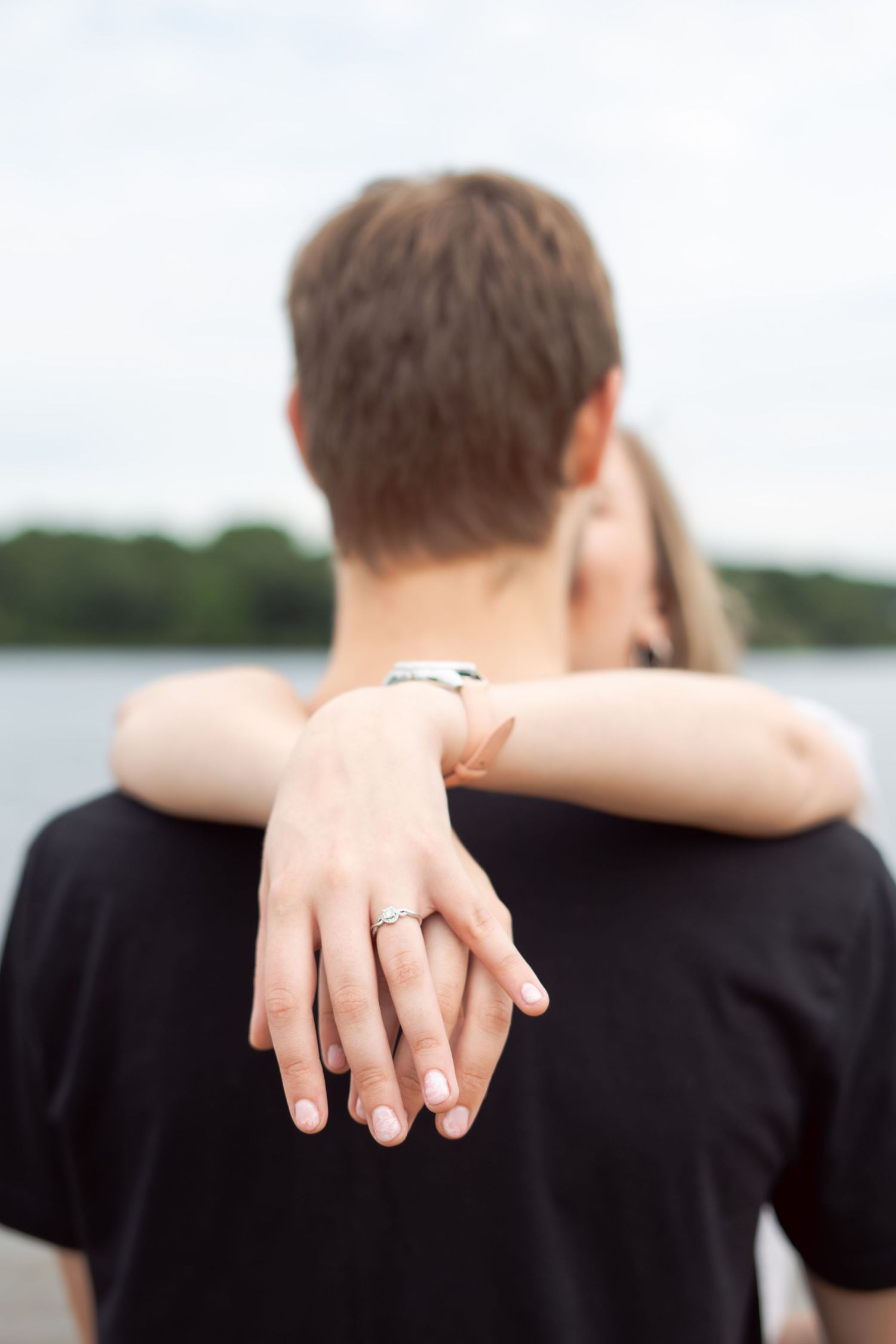 Close-up of hands embracing showing an engagement ring against a waterfront backdrop.