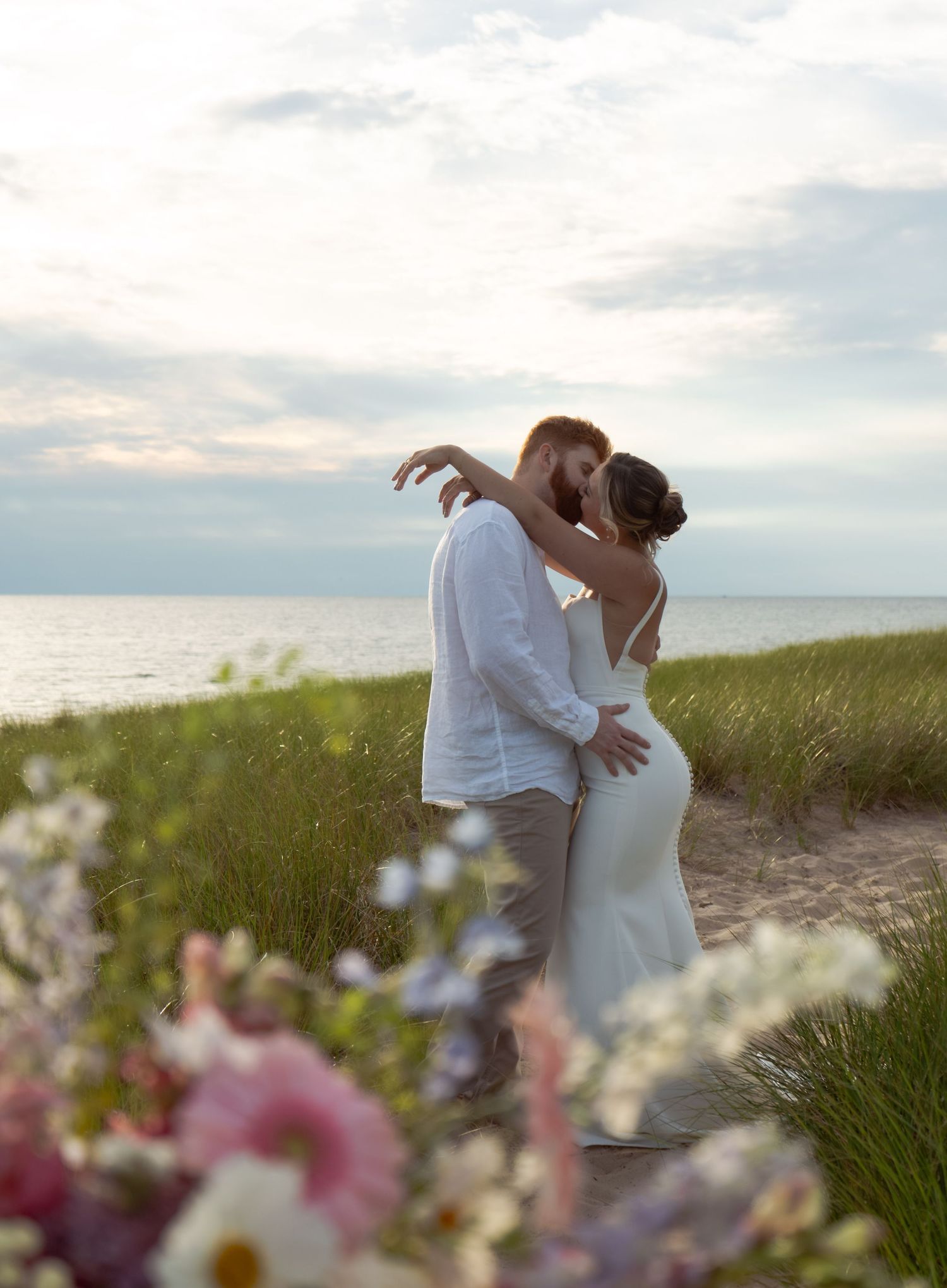 A romantic beachside moment between two figures embracing at sunset with wildflowers in the foreground.