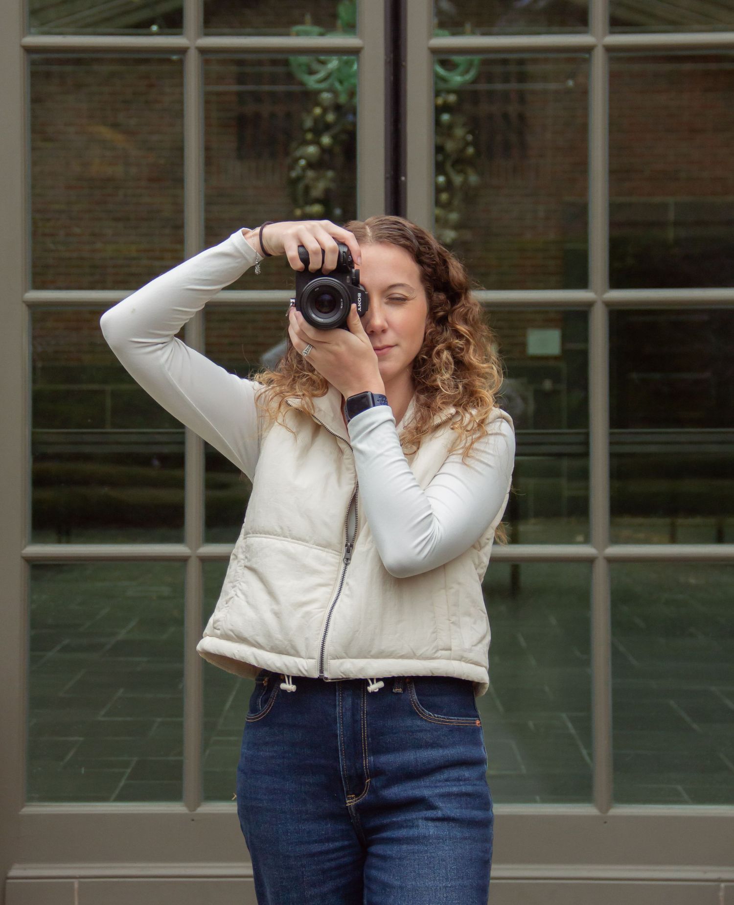 Photographer in a white jacket and jeans taking pictures with a professional camera near a windowed wall.