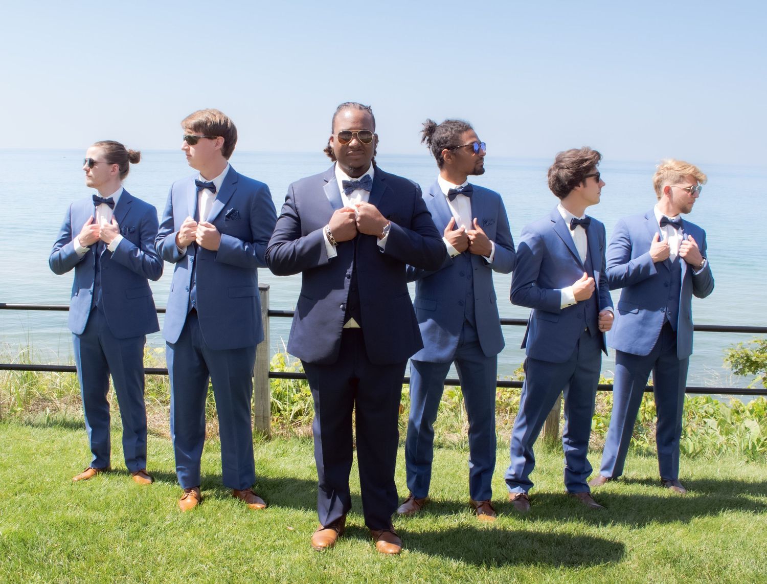 Wedding party in matching navy suits stands in a line outdoors adjusting their ties on a sunny day.