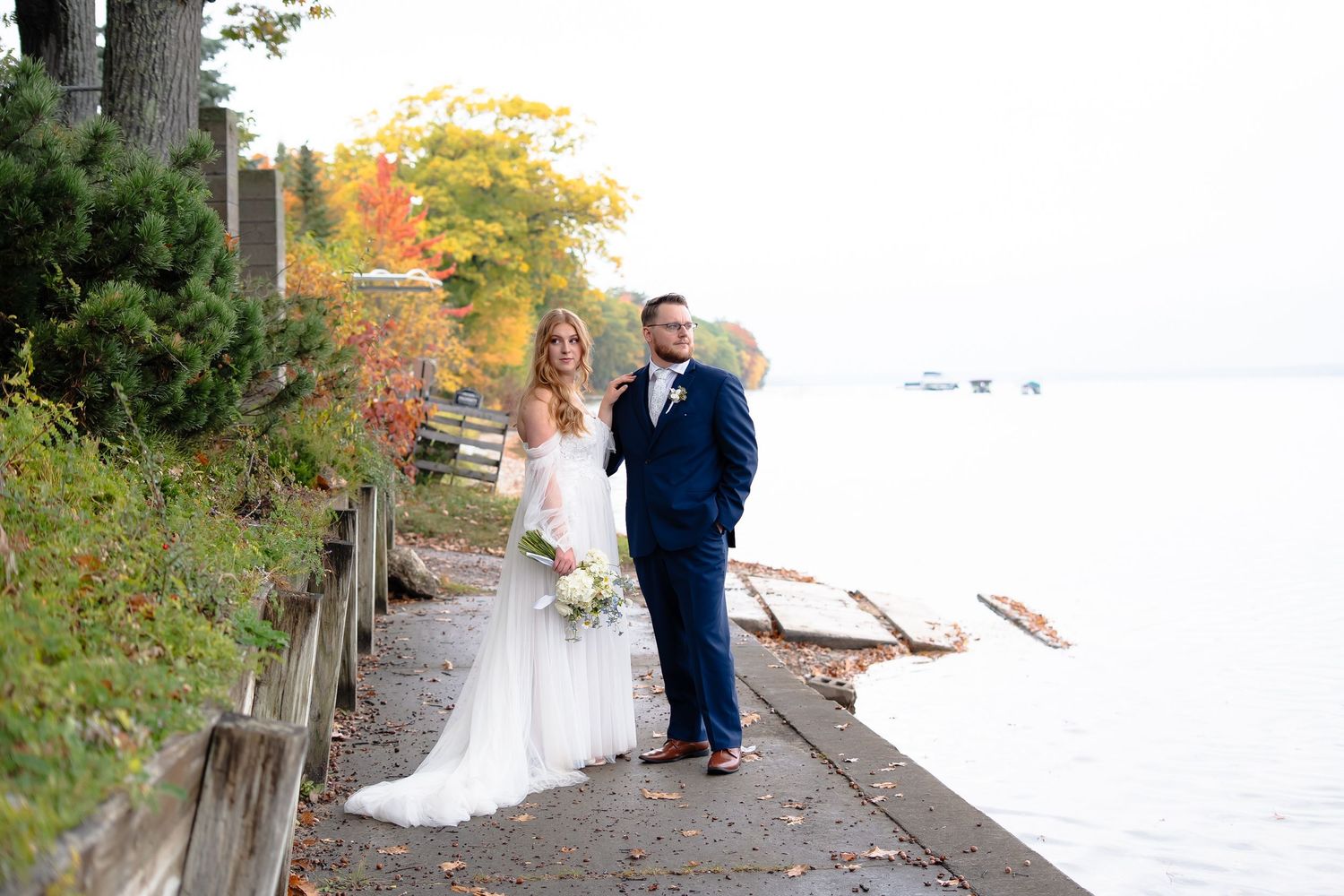 Couple in wedding attire walking along a wooden boardwalk surrounded by autumn foliage and water views.