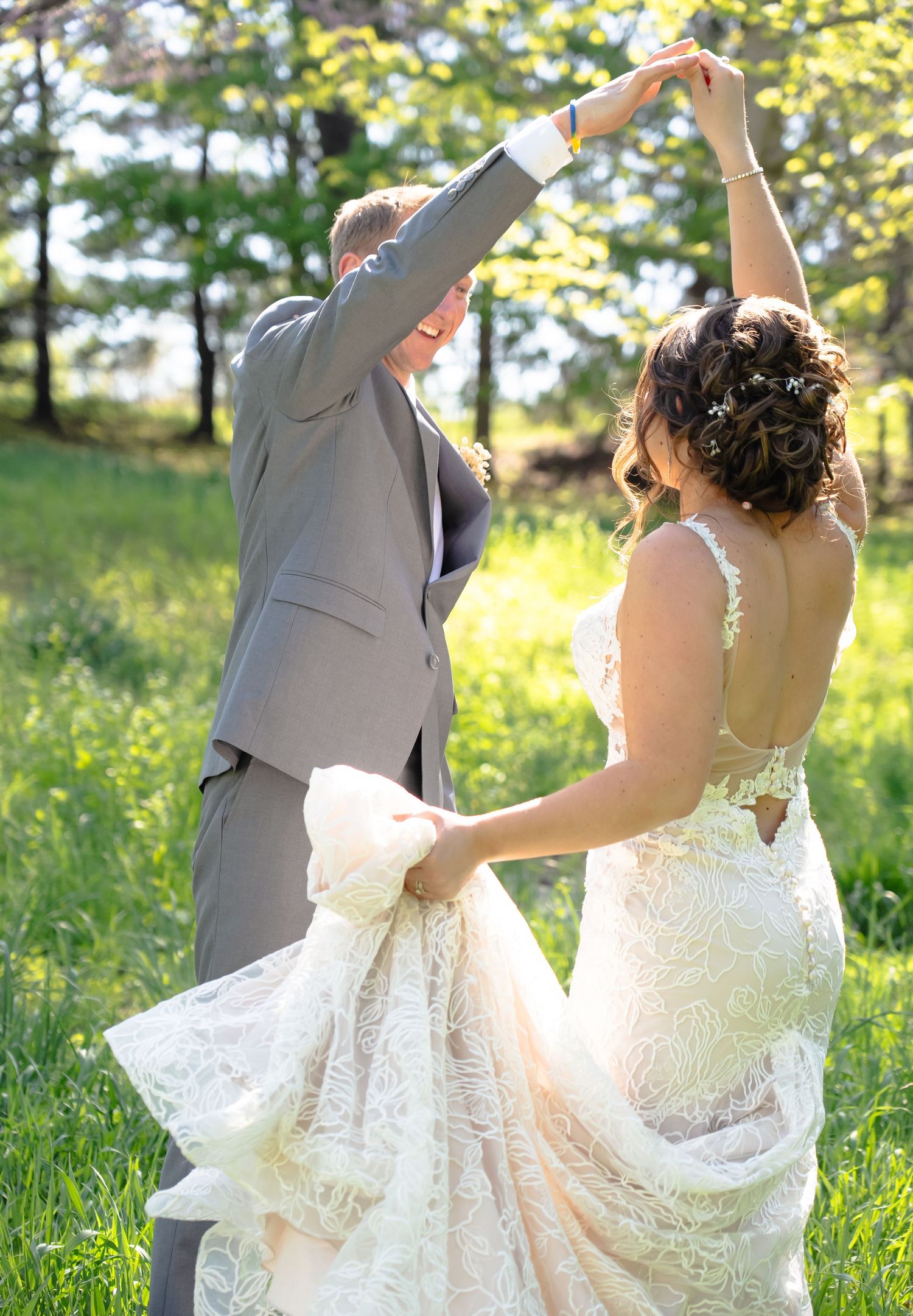 Newlyweds dancing together in a sunlit meadow with trees in background as bride's lace dress flows gracefully.