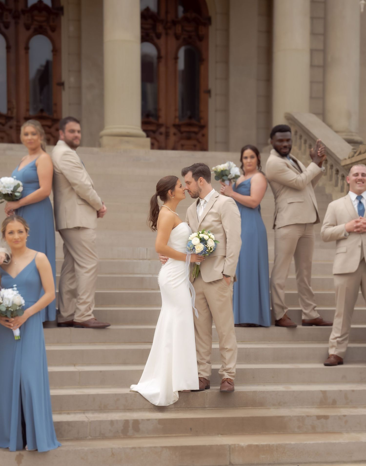 A wedding party poses on grand stone steps with bridesmaids in blue dresses and groomsmen in beige suits.