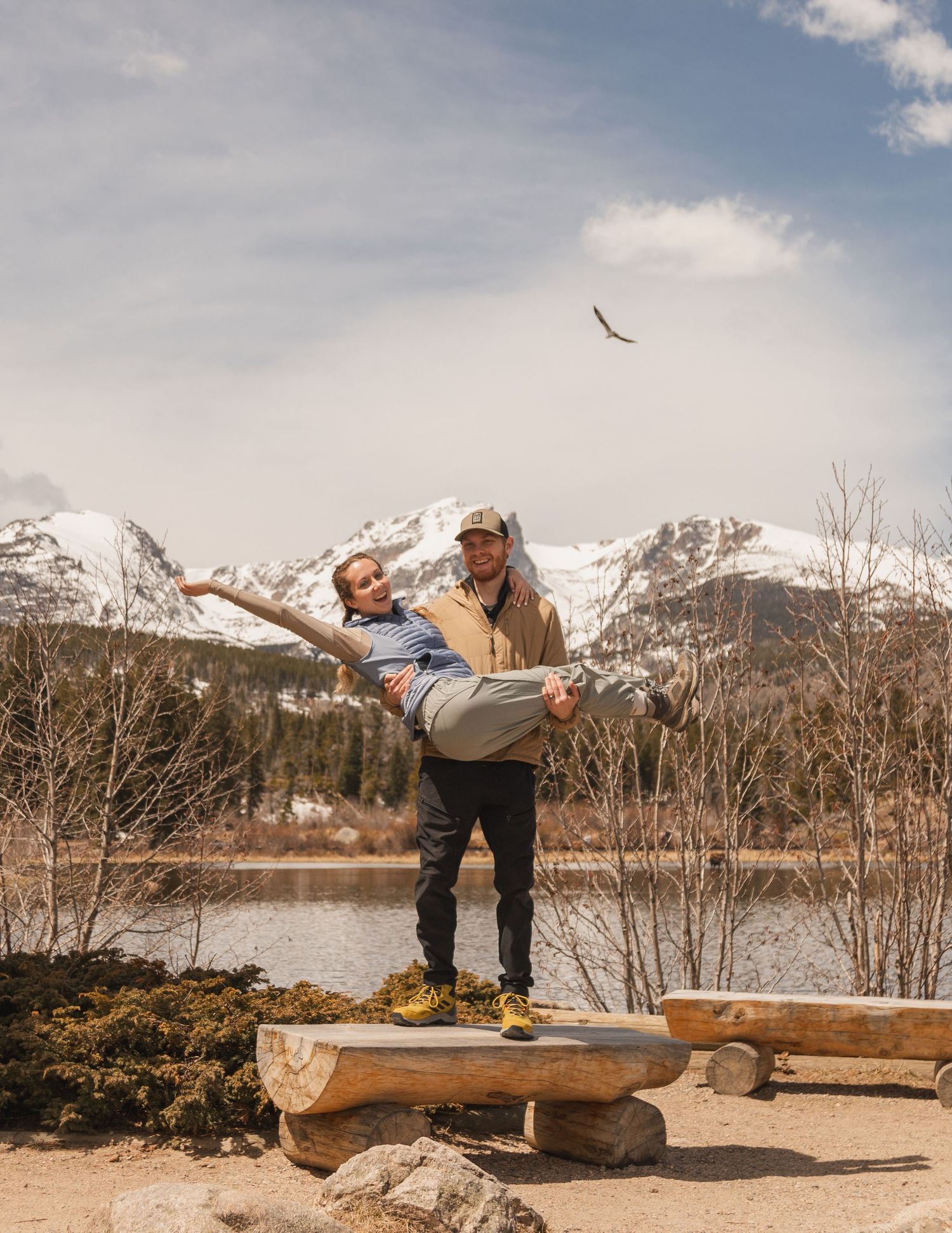 A playful couple poses against snowy mountain backdrop with a soaring bird overhead.
