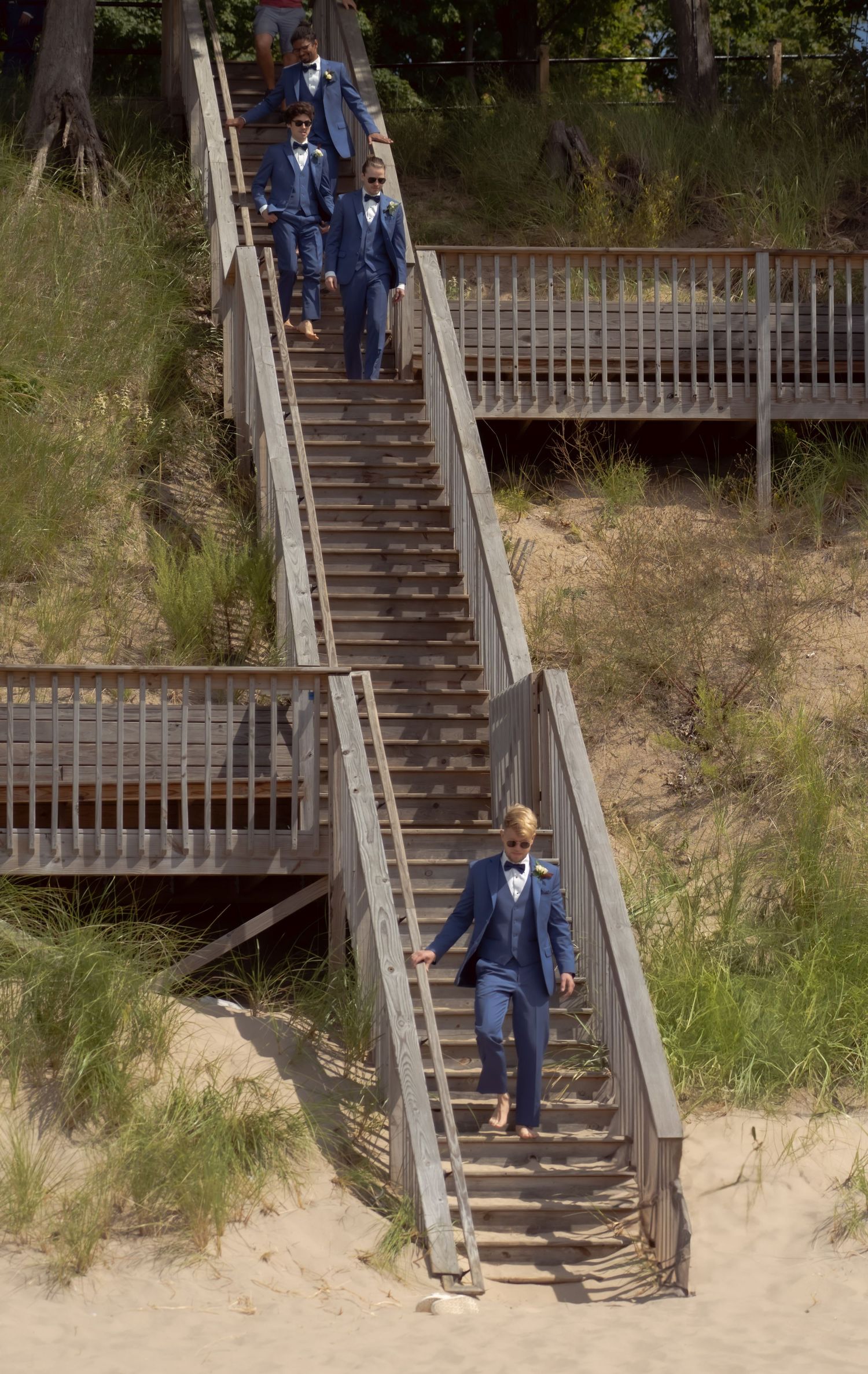 Groomsmen descend a wooden staircase on a sandy hillside wearing matching navy blue suits.