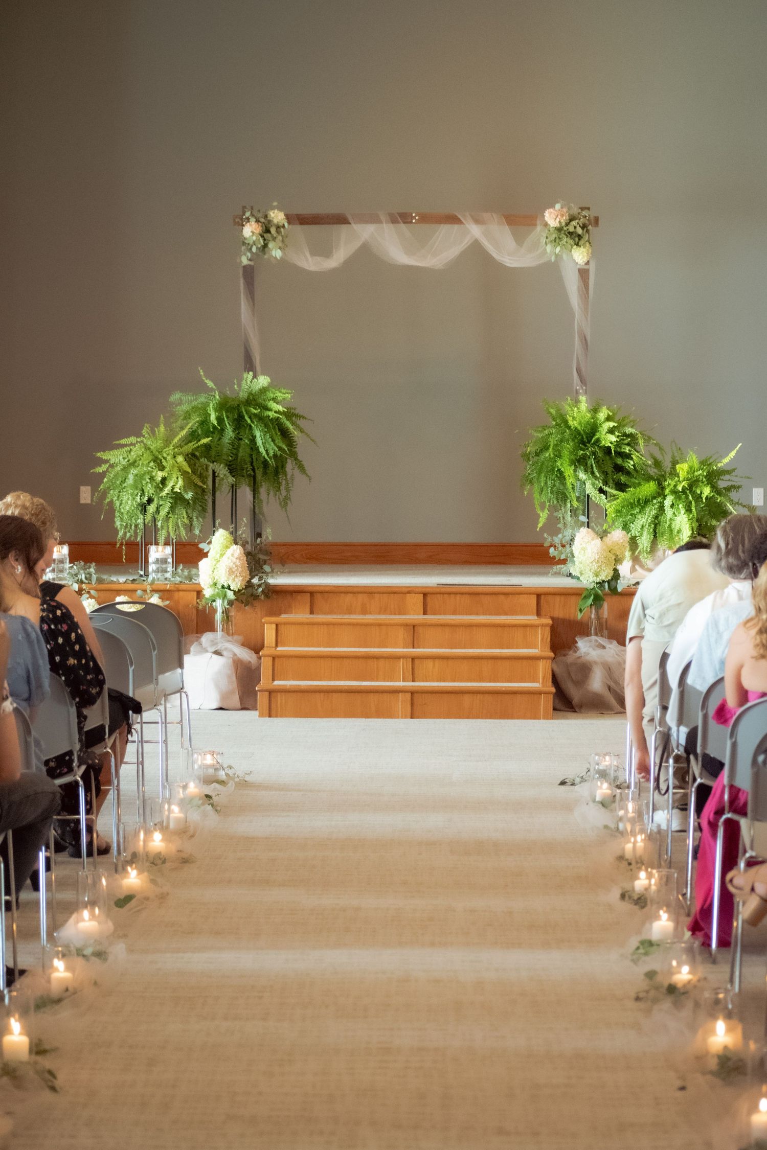 Wedding ceremony setup with fern-decorated arch, wooden steps, and candle-lined aisle in an indoor venue.