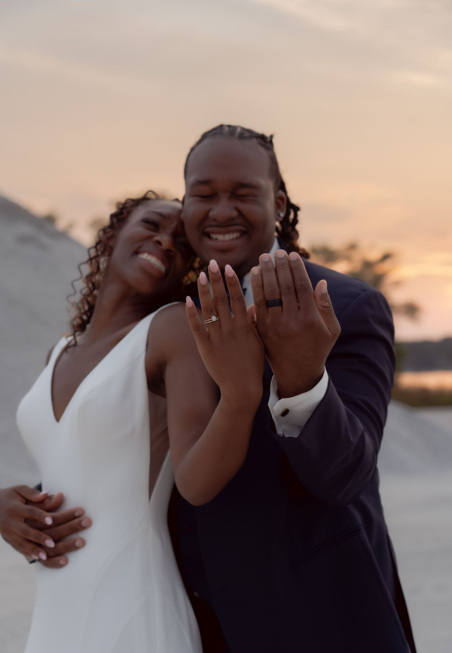 A joyful couple laughs together at sunset while showing off their wedding rings in a candid moment.