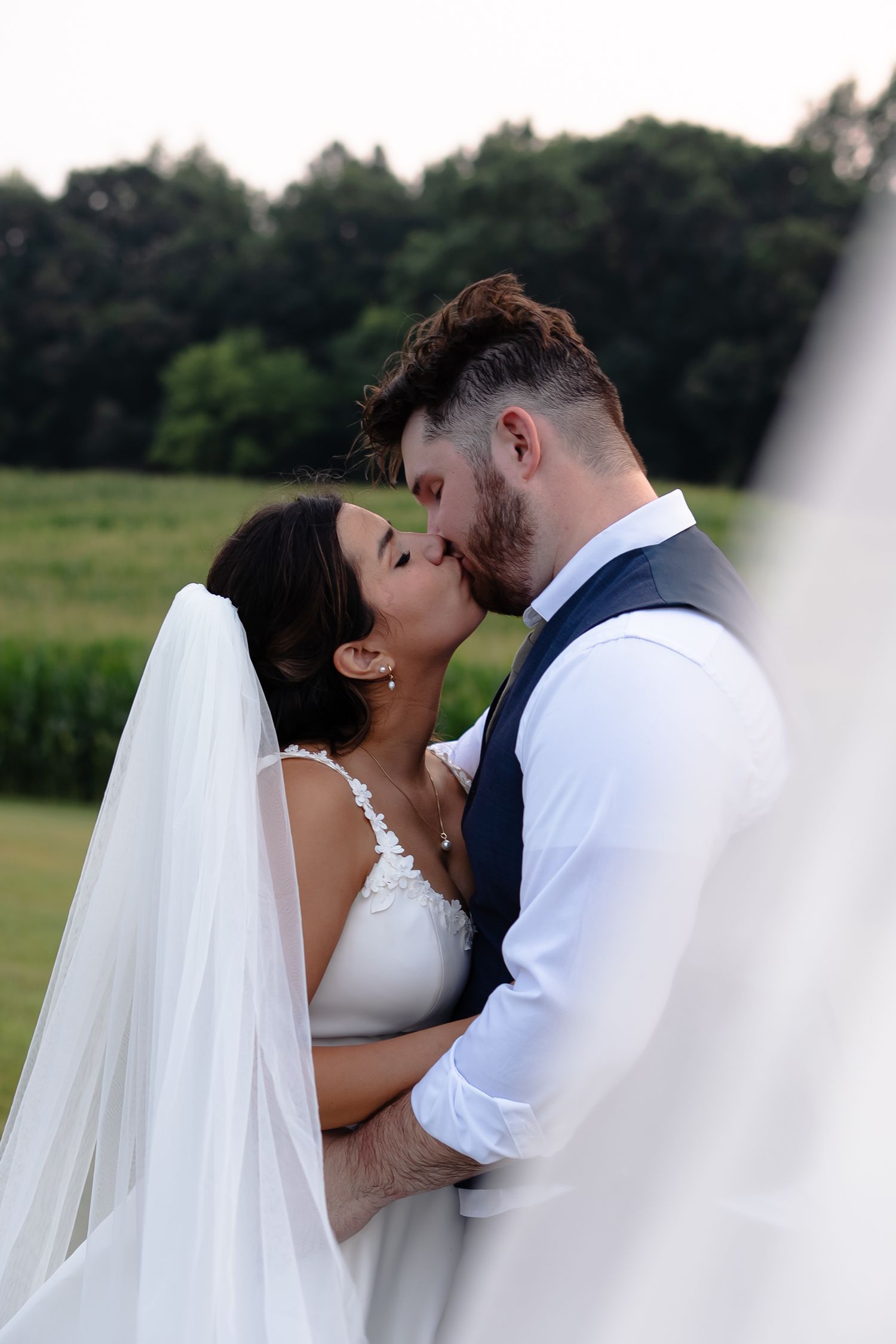 A romantic kiss between newlyweds outdoors with the bride in a white gown and veil and groom in vest and shirt.