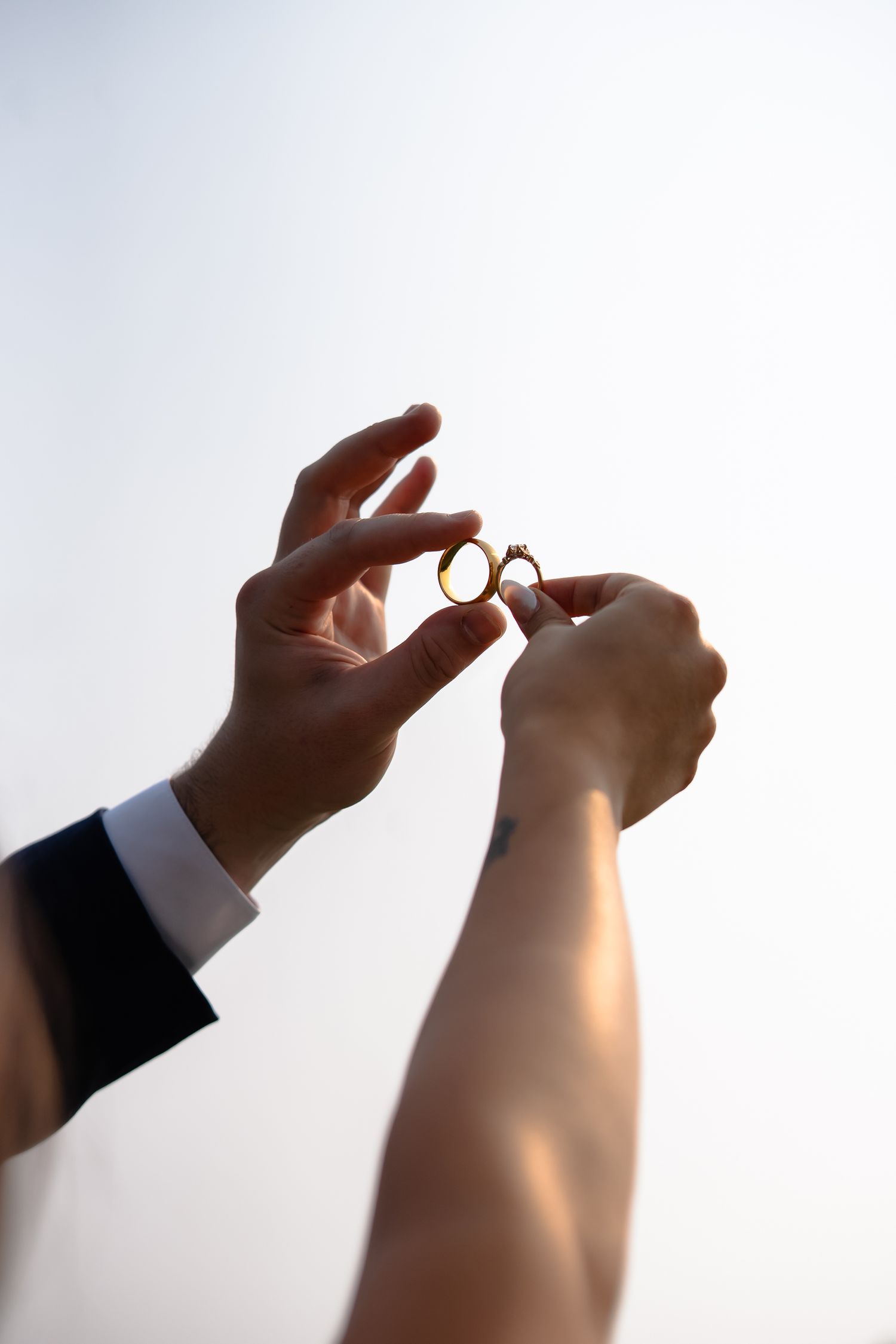 Hands in formal attire hold up wedding rings against a bright sky background in silhouette.